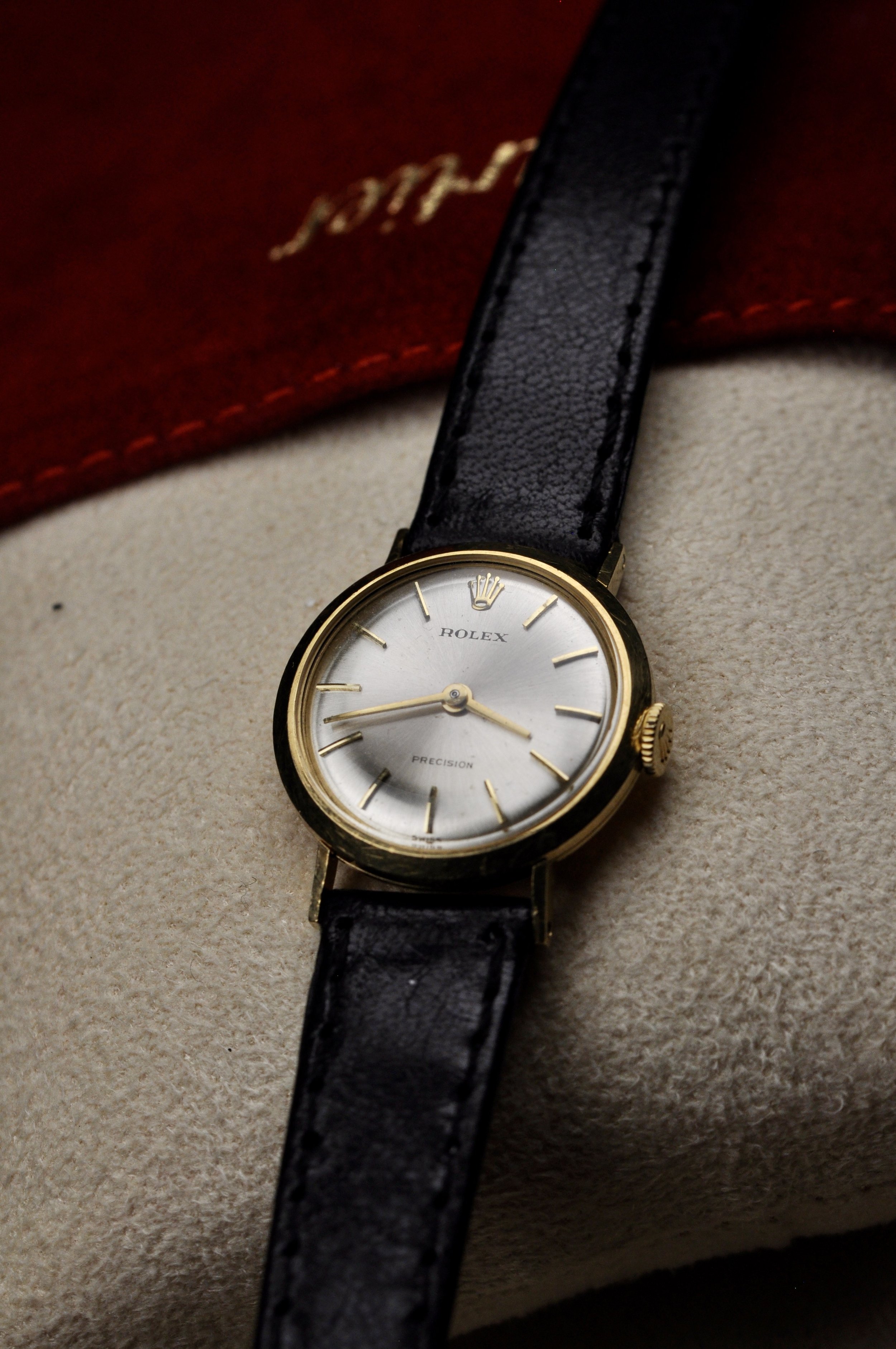 A vintage Rolex wristwatch with a gold case, white dial, and black leather strap is displayed on a beige surface with a red box in the background.