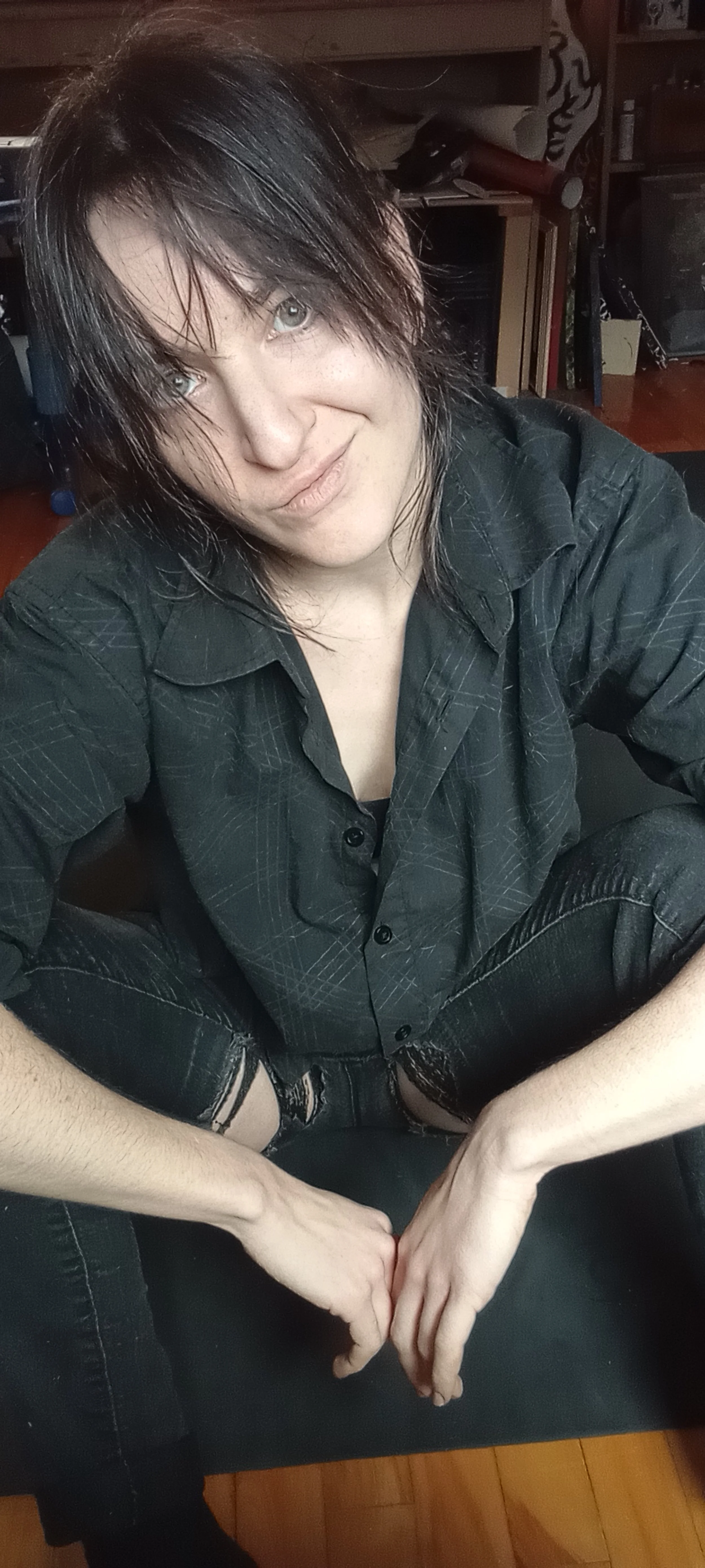 Une personne à cheveux noirs et yeux clairs, souriant et vêtue d'une chemise noire, est assise sur un canapé en bois dans une pièce avec des étagères.