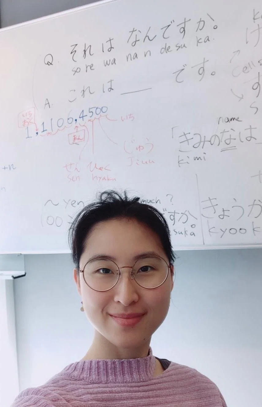 A woman with short black hair and glasses smiling in front of a whiteboard filled with Japanese writing and math notes.