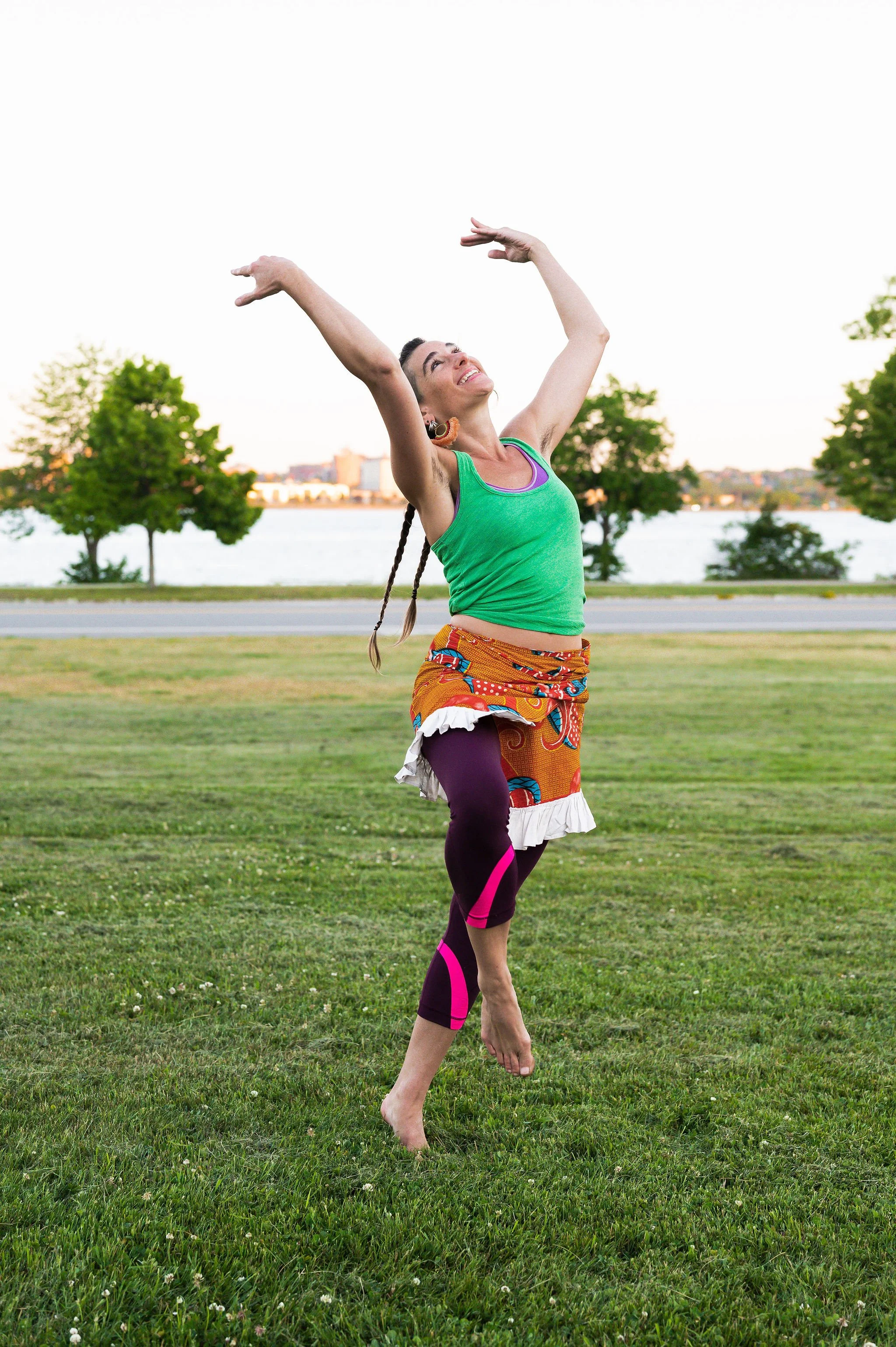 Marita has two long braids and is dancing in a field with a view of an ocean cove in the background. She is wearing a bright green tanktop, patterned orange skirt and purple leggings and is gesturing towards the sky, which she's also looking up at.