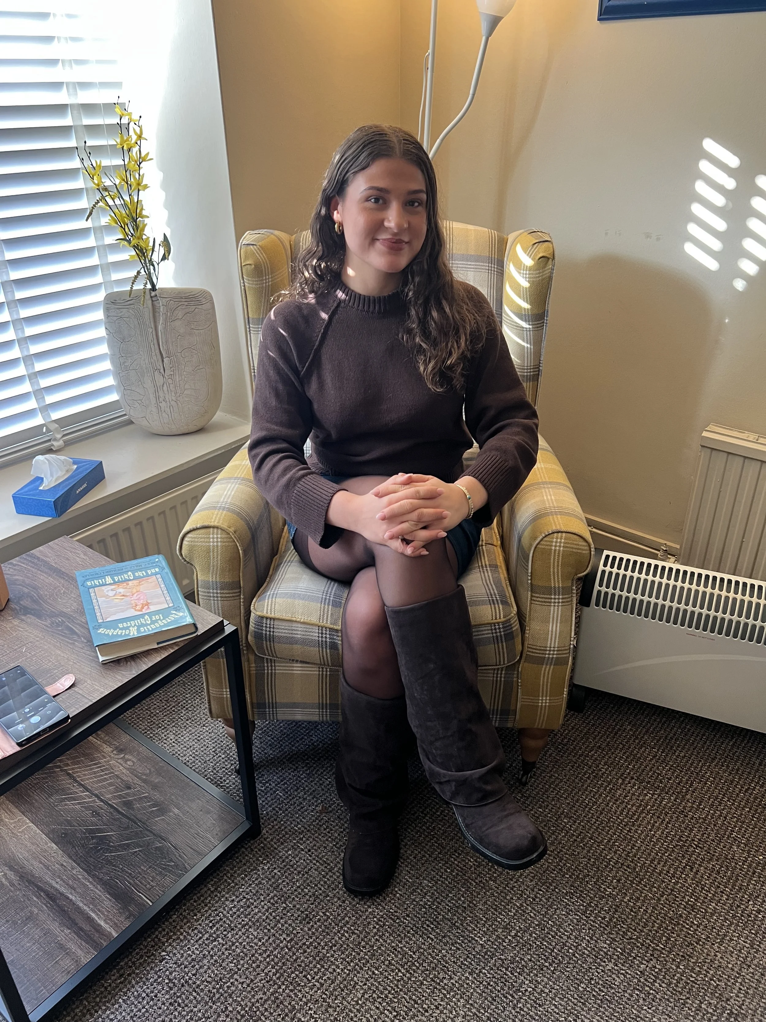 A woman sitting on a yellow checkered armchair in a cozy room, with a window, a white vase with yellow flowers, and a small table holding a book, tissue box, and a phone.