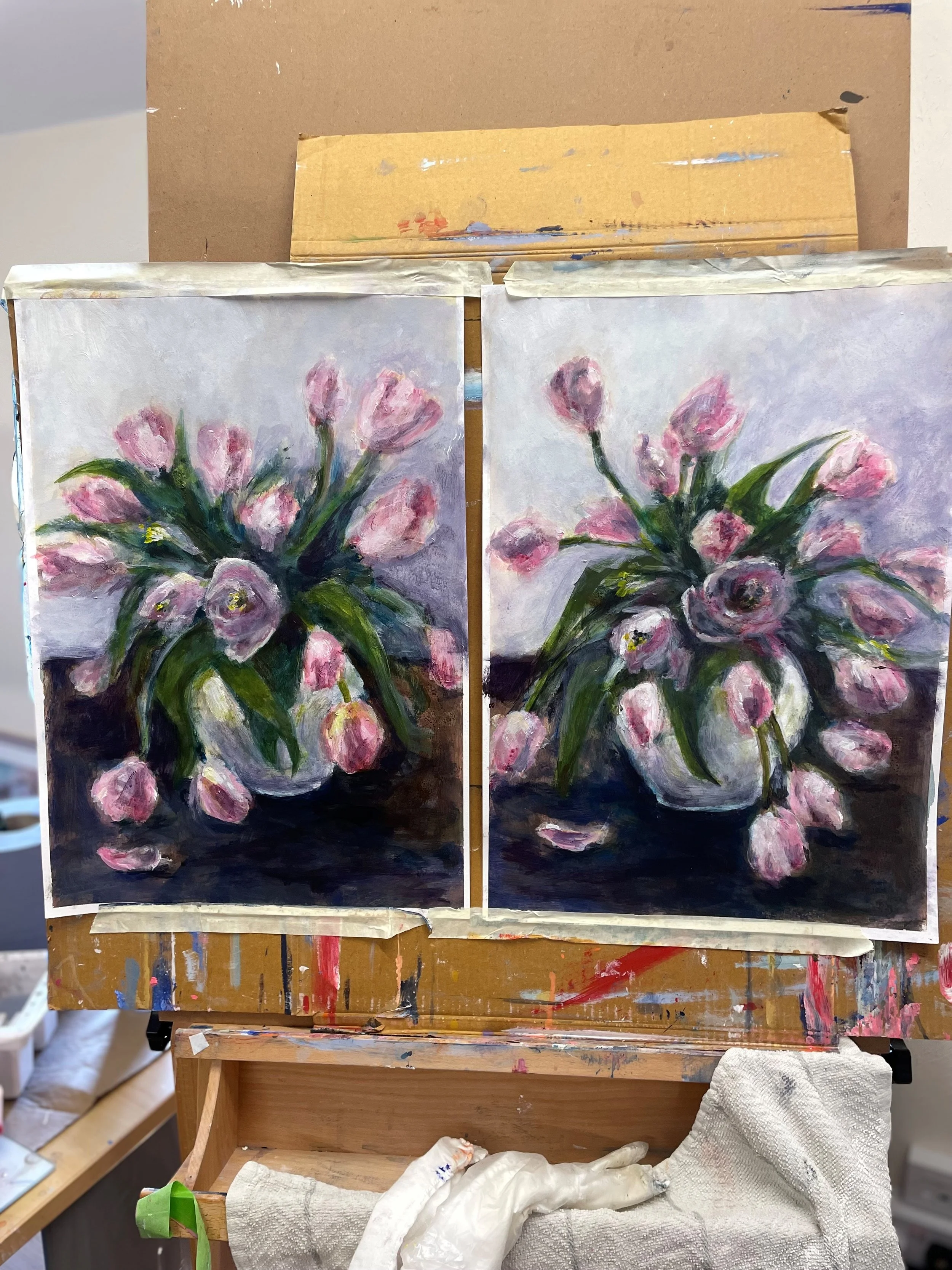 Two small canvas paintings of pink tulips with green leaves in a vase, set on a wooden table with art supplies and a white cloth.
