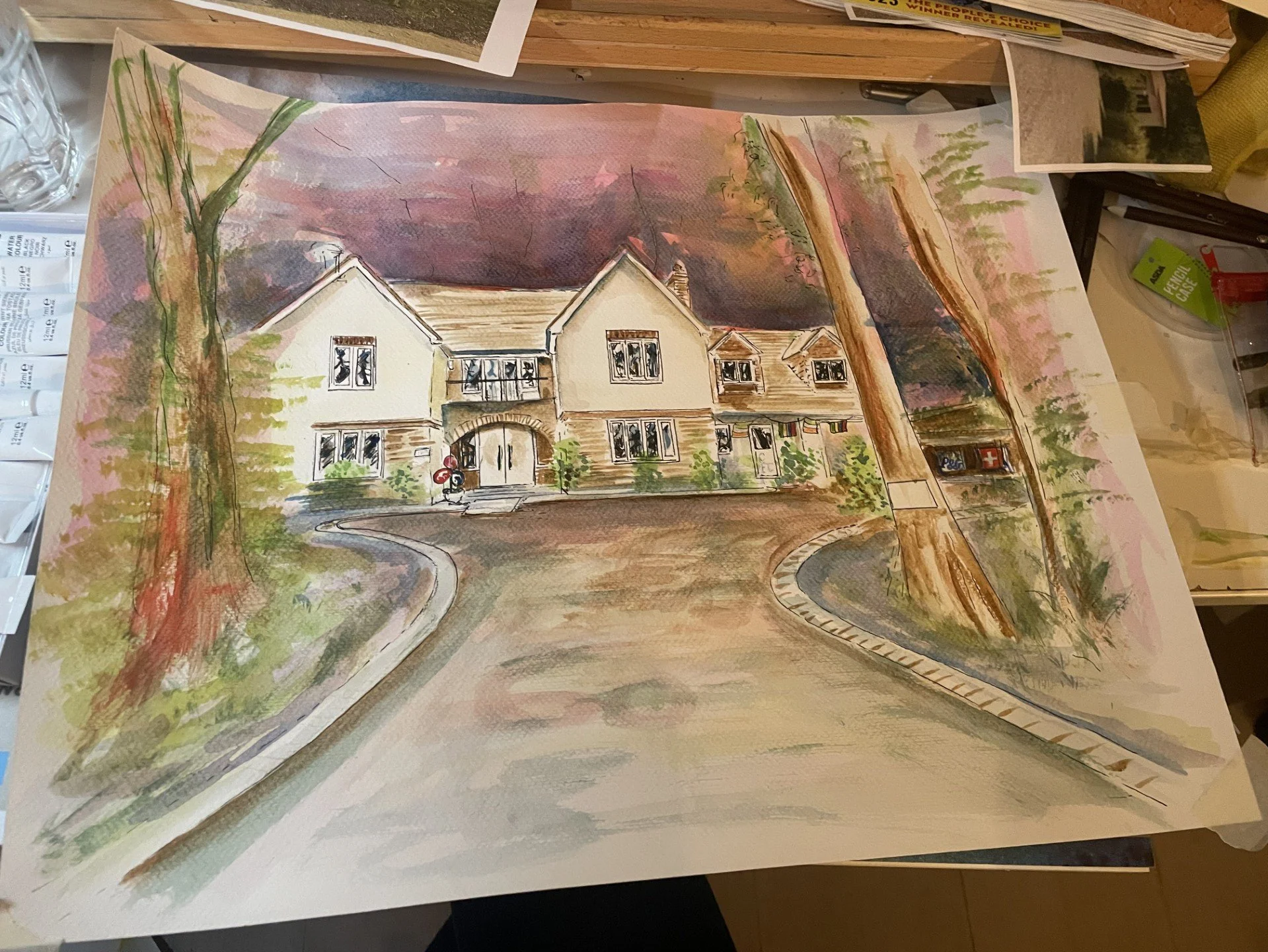 A watercolor painting of a house with trees and a driveway, with some newspapers and art supplies scattered around.