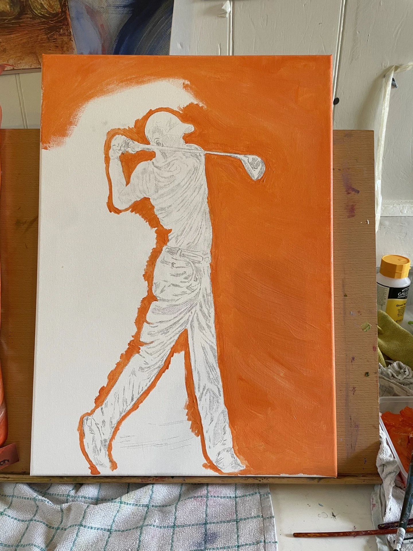 A painting in progress of a golfer preparing to swing, with the figure outlined in white against an orange background. The artist's workspace with painting tools and a towel is visible.