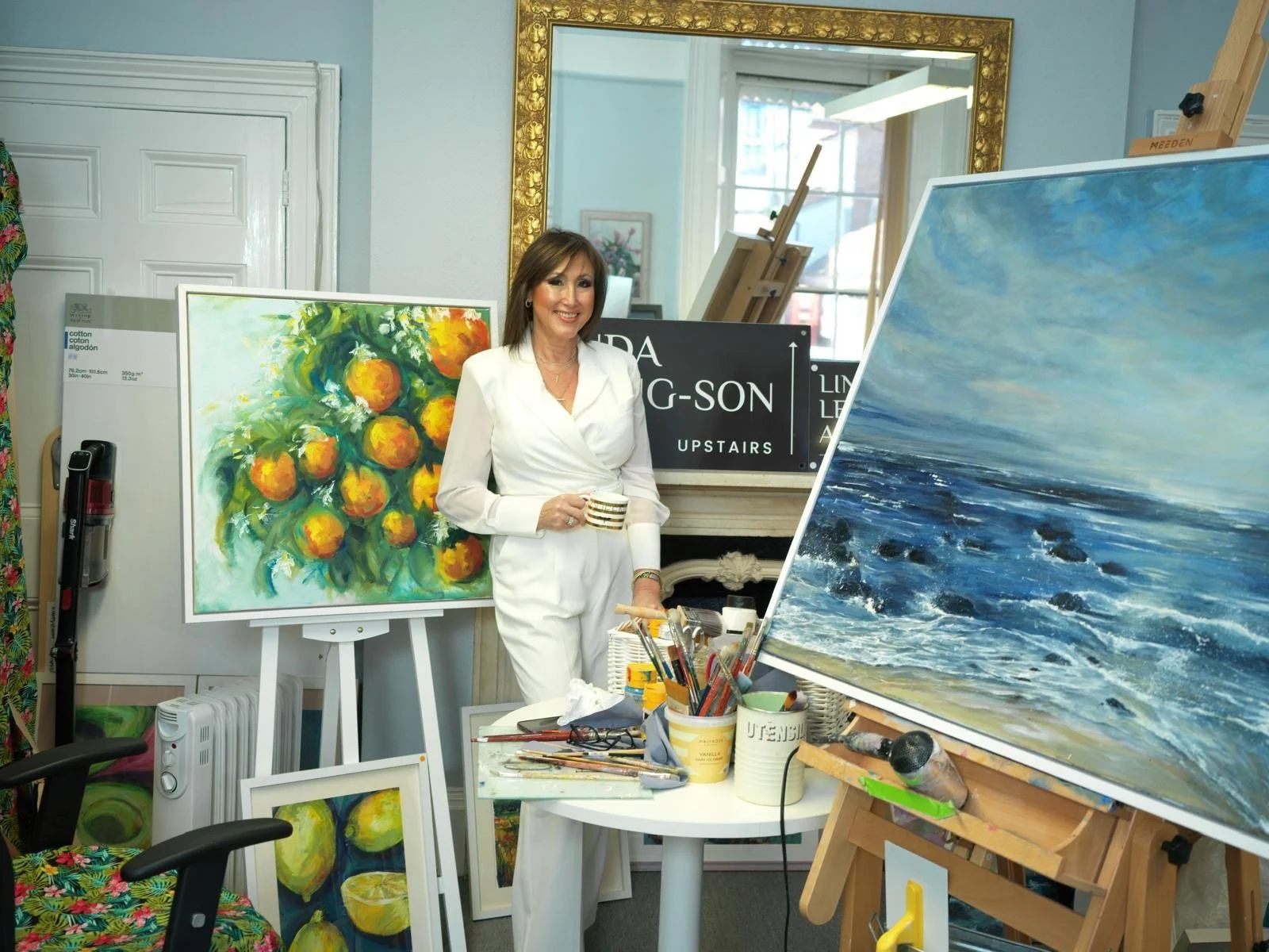 A woman in a white outfit standing in an art studio, holding a coffee mug. Behind her are colorful paintings, including a still life of oranges and a seascape with blue waves. There are art supplies on a table in front of her.