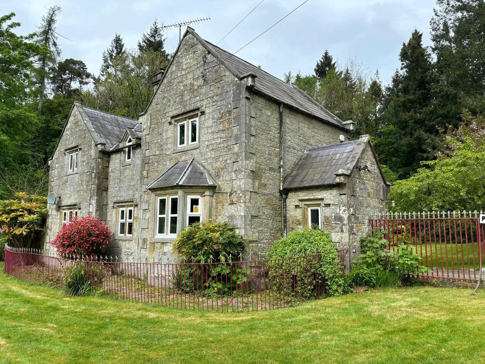 A large stone house surrounded by a garden with various shrubs and flowers, enclosed by a red metal fence. The house has multiple windows and a steeply pitched roof, set against a backdrop of tall trees and an overcast sky.
