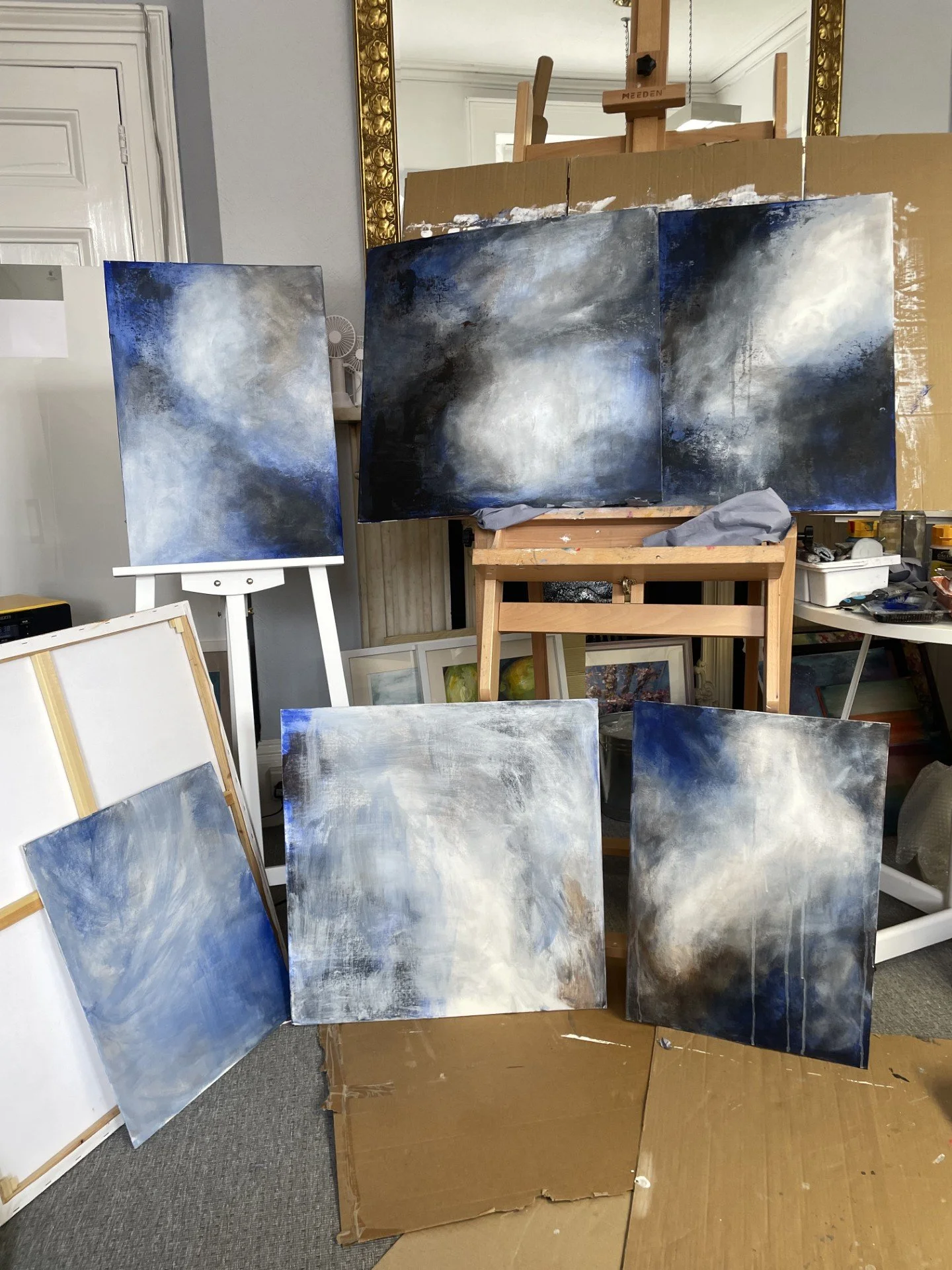 Several abstract landscape paintings with blue, white, and dark tones are displayed on and around a cardboard and easel in an art studio.