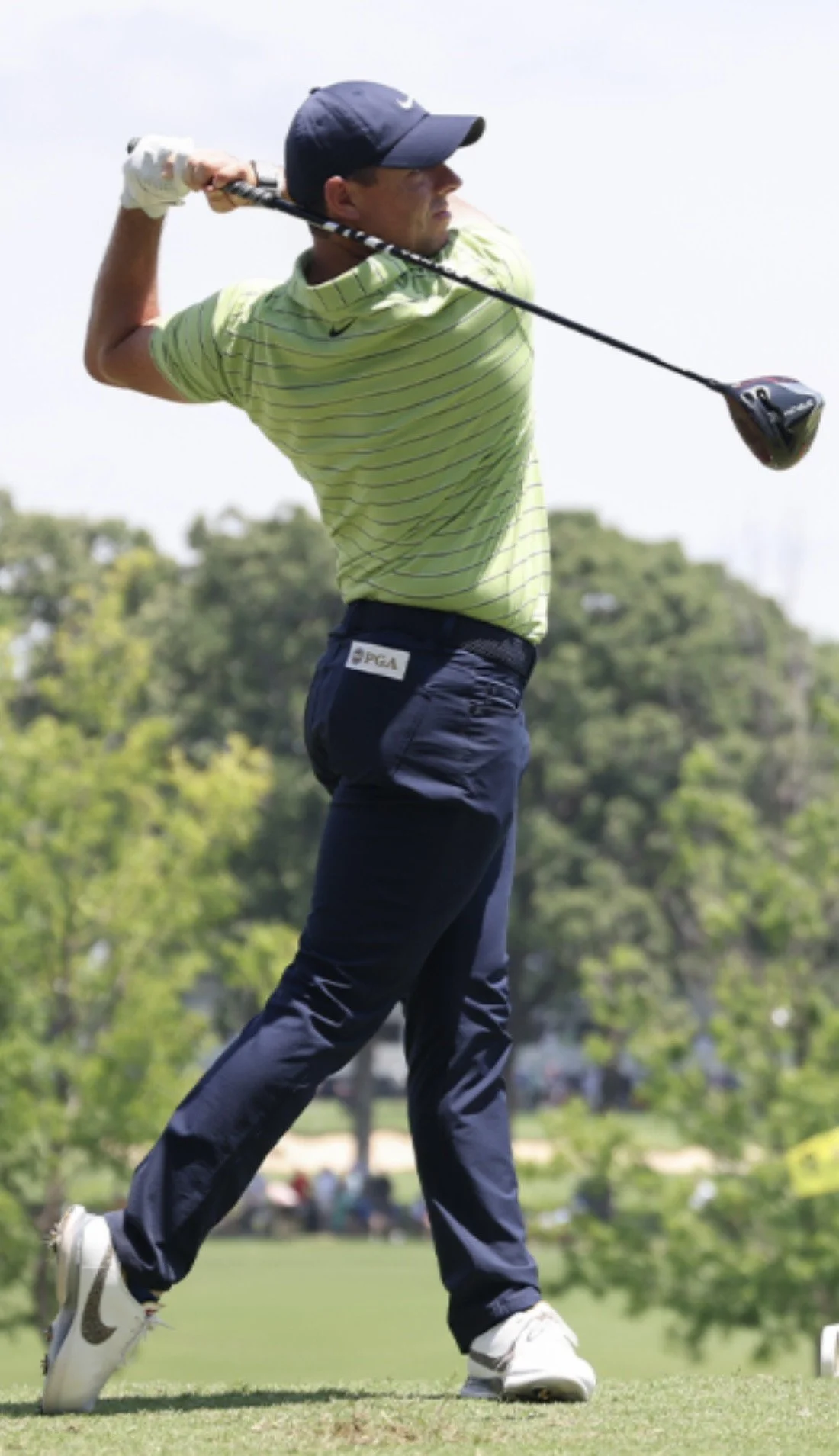 A male golfer in a green striped shirt and navy pants swinging a golf club on a golf course with trees in the background.