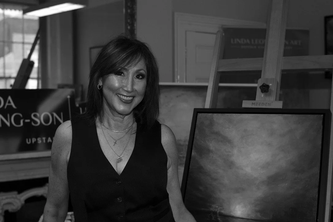 A woman smiling in an art gallery or studio, surrounded by framed paintings and artwork.