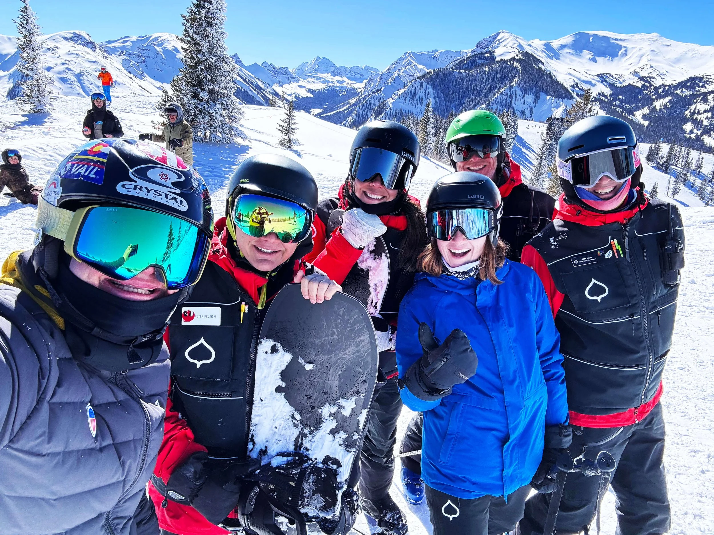 Group of six people in ski gear smiling on snowy mountain with ski slopes and mountains in the background.