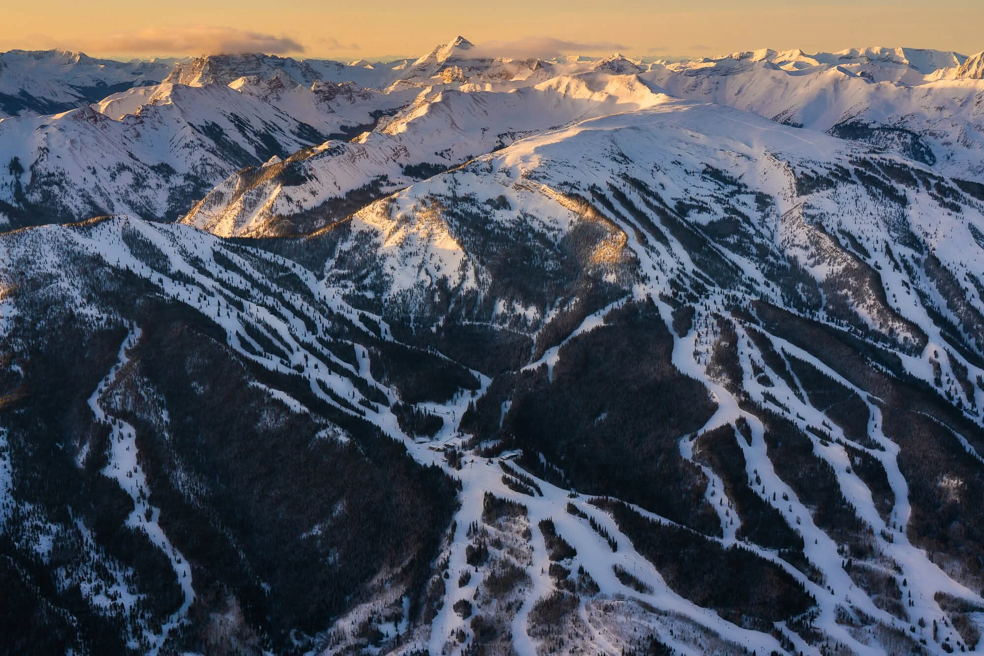Snow-covered mountains with ski slopes and dark forests in the foreground, under a partly cloudy sky at sunset.