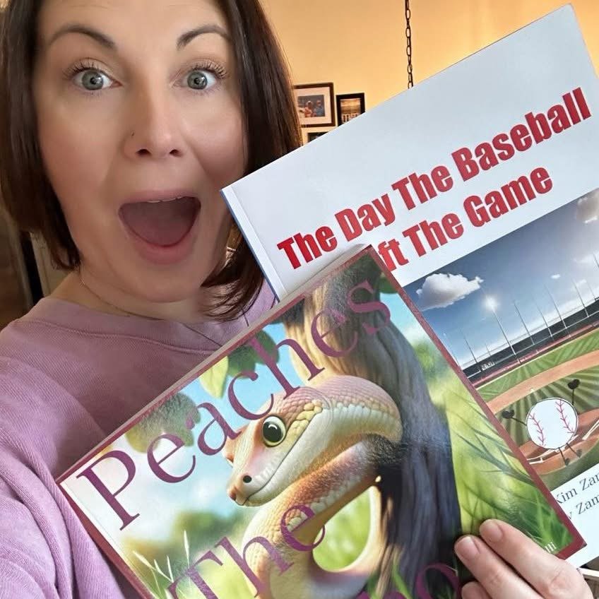 A woman with brown hair and a pink shirt is holding two books and has an excited expression. One book is titled "The Day The Baseball Hit The Game" and the other is a children's book titled "Peacocks" featuring a snake with a peacock's head.