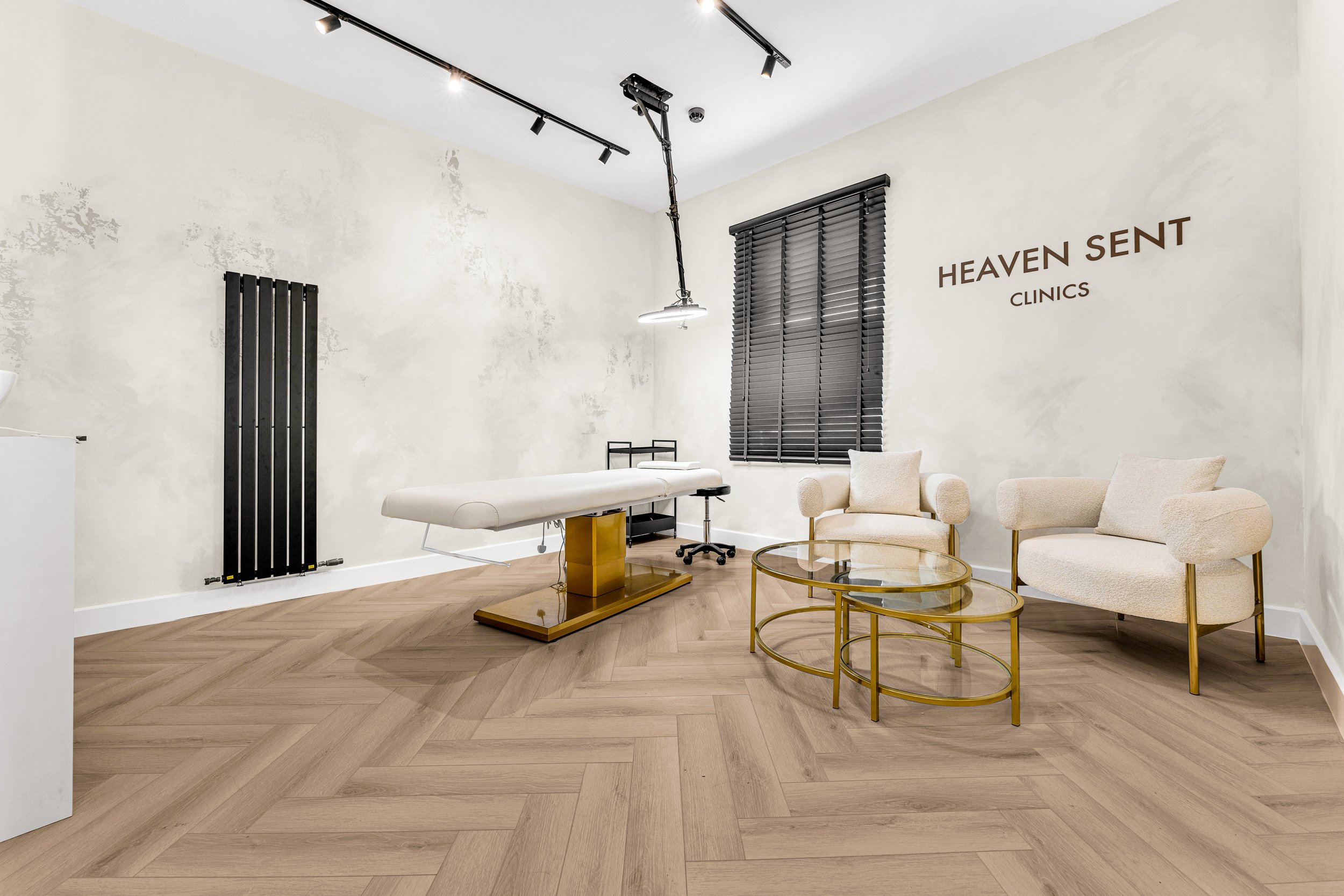 A modern, minimal medical clinic waiting area with two light-colored armchairs, a clear glass coffee table with gold frame, a massage or examination table, black window blinds, a black wall radiator, and a sign on the wall that reads 'HEAVEN SENT CLINICS'.
