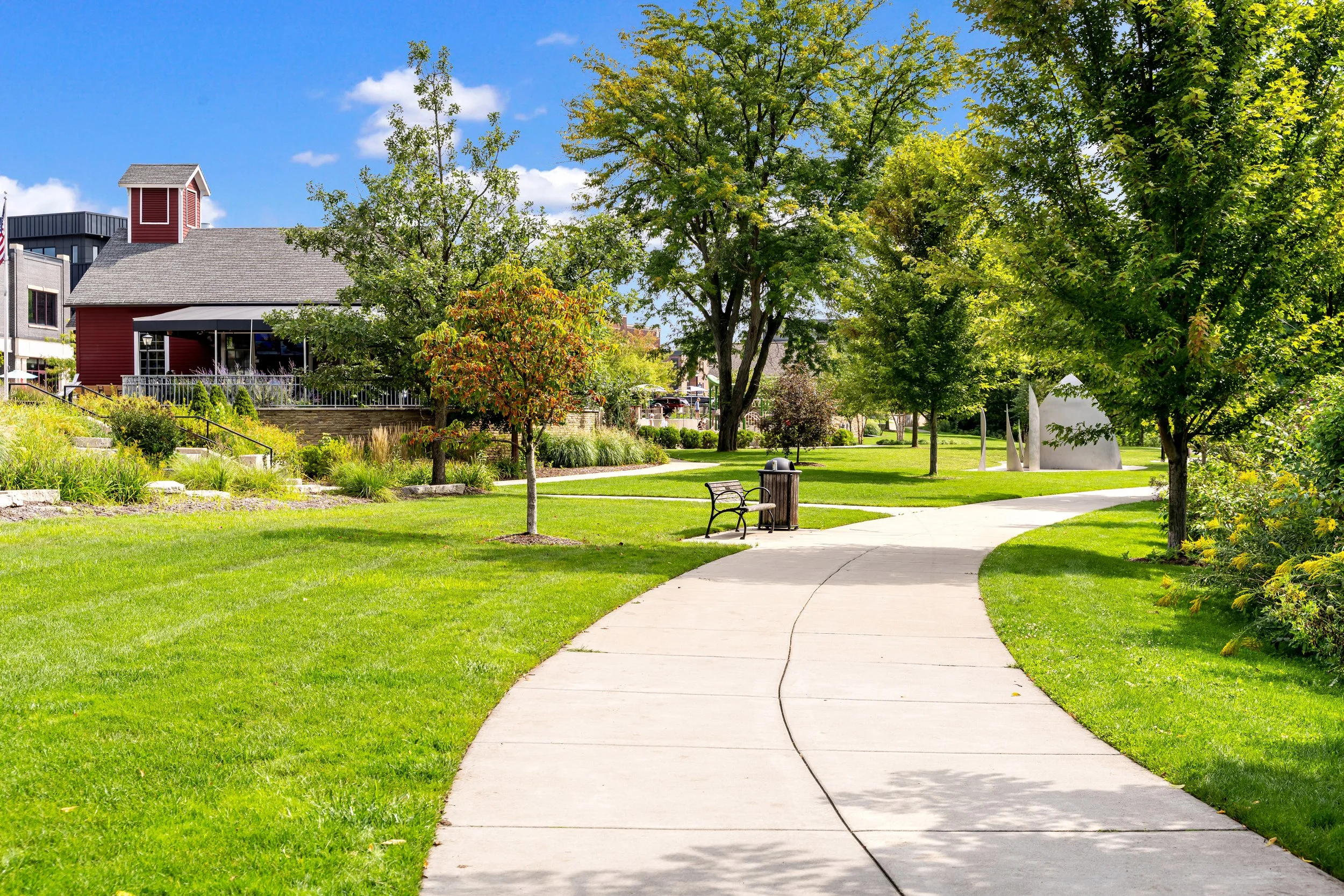 A sunny park scene with a curved concrete path, green grass, trees with lush foliage, a bench, and a trash can, with buildings and a blue sky in the background.