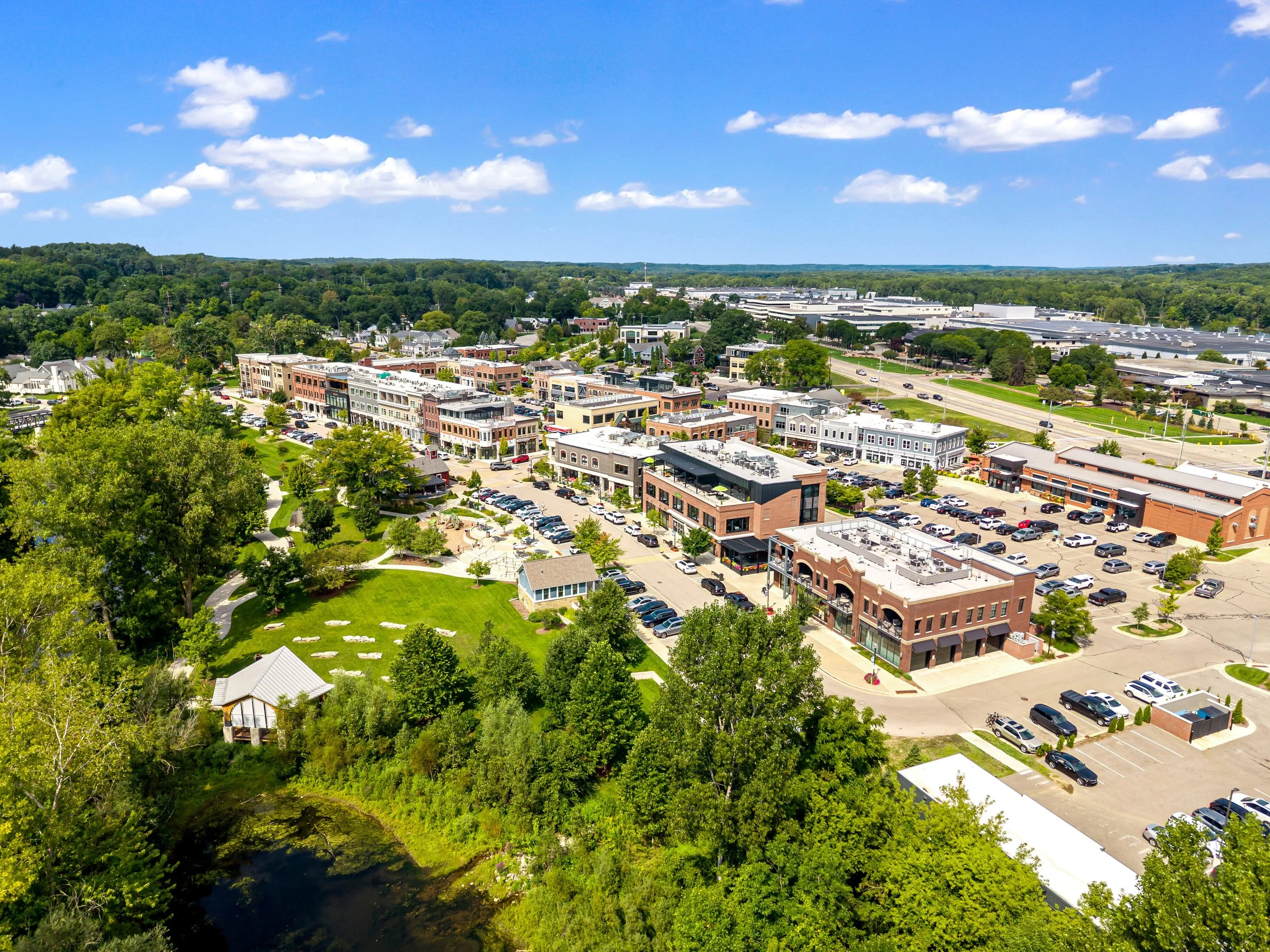Ada Michigan West Michigan Aerial view of a small town with colorful buildings, green trees, a parking lot, with a blue sky and scattered clouds.