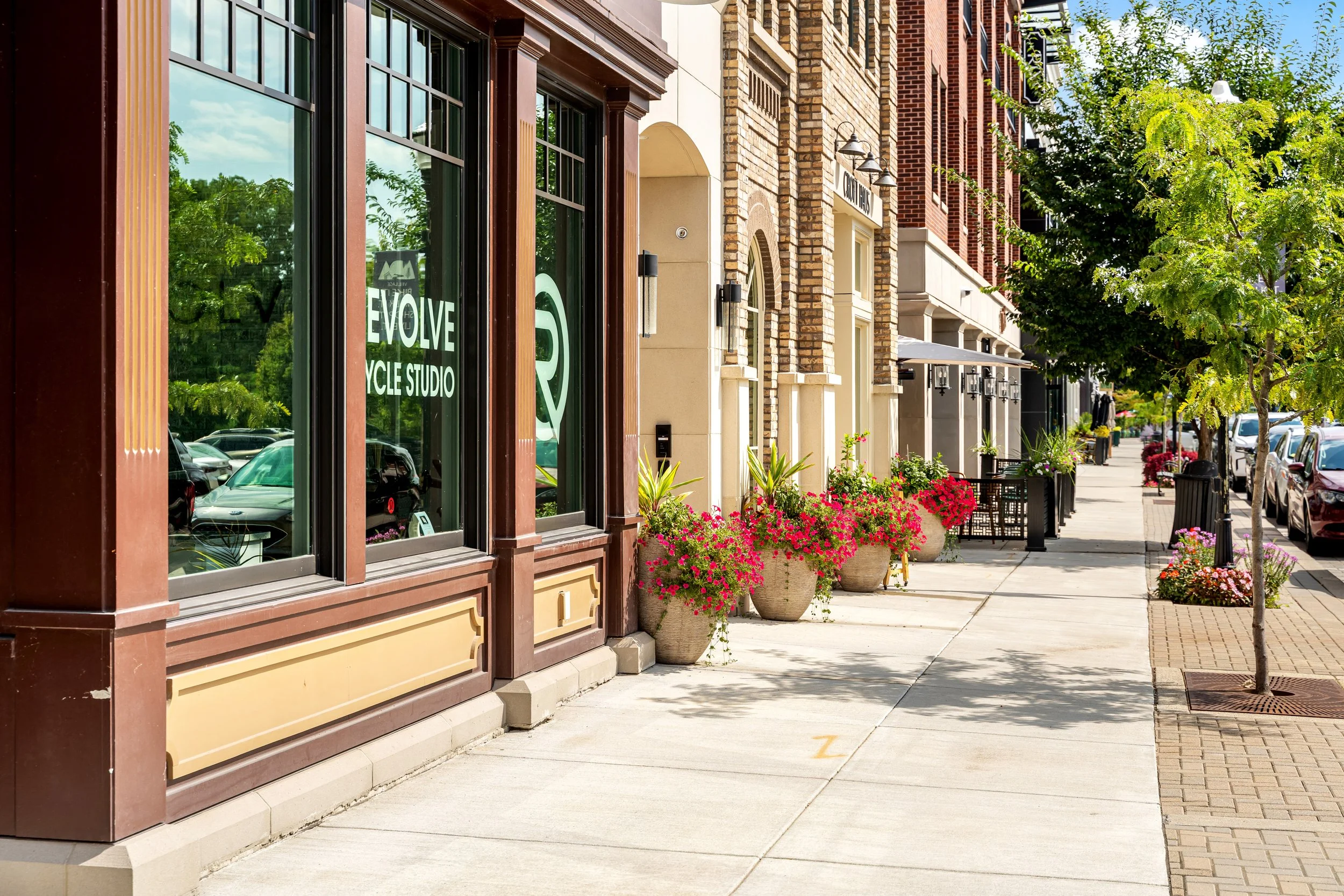 A sidewalk lined with shops and storefronts with large windows, trees, and colorful flowers in pots. There are parked cars on the street and a clear blue sky overhead.