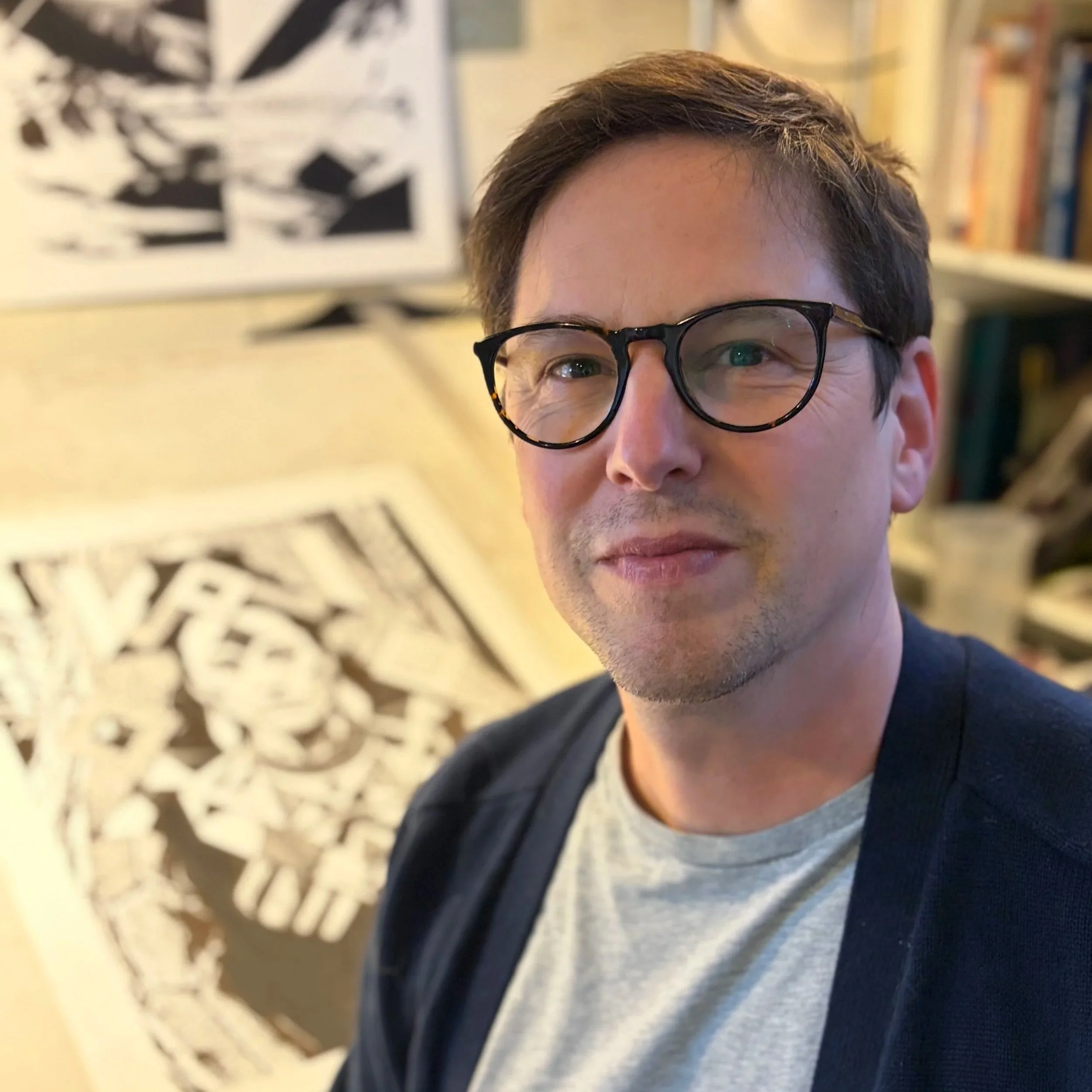 A man with dark hair, glasses, and a light beard taking a selfie in an art studio, with black and white artwork in the background.