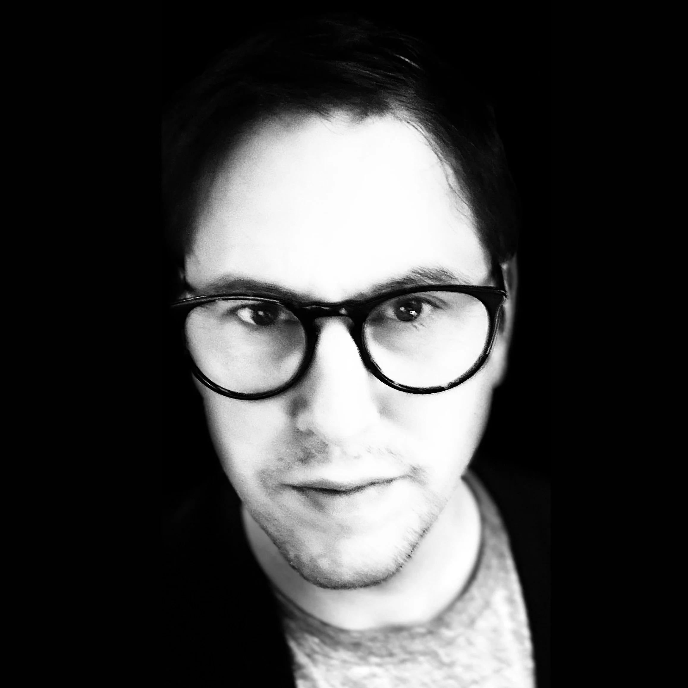 Black and white close-up portrait of a man wearing large round glasses, looking directly at the camera, with dark hair and a slight smile.