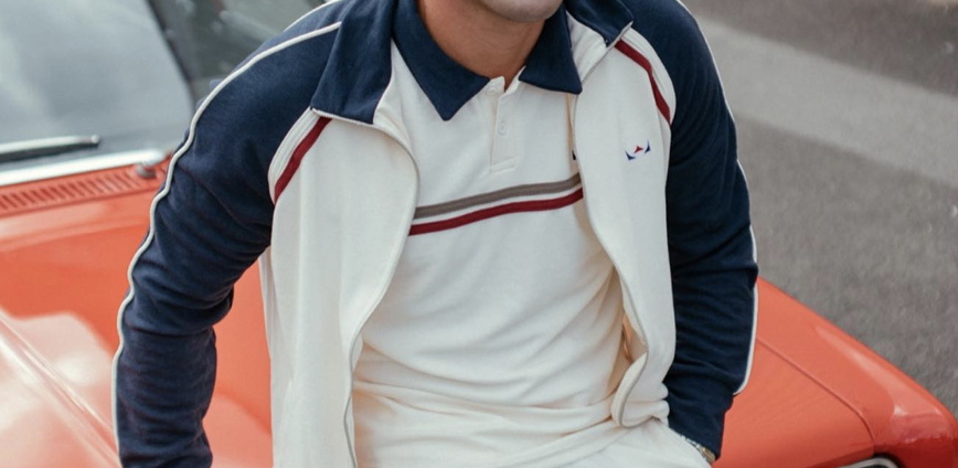 Close-up of a person leaning on a red car, wearing a white polo shirt with red and navy stripes and a navy and white jacket.