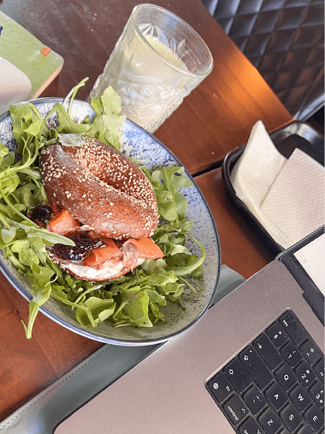 A bowl of salad with a sesame-seeded bagel sandwich on top, placed on a wooden table with a glass of water and a laptop nearby.