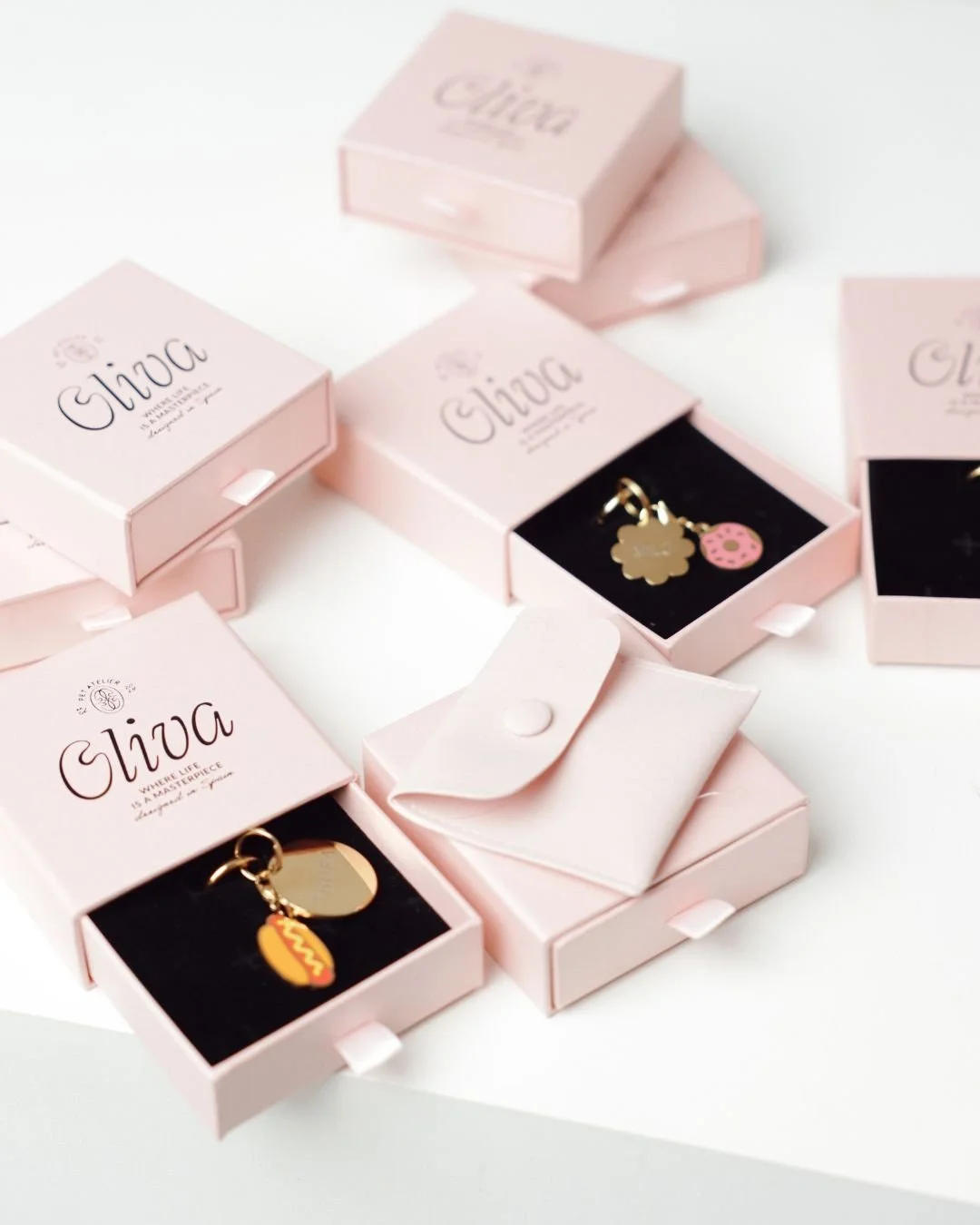 Several pink jewelry boxes with the brand name 'Oliva' are arranged on a white surface. One box is open, revealing a gold charm with a hotdog pendant and a small pink donut charm.