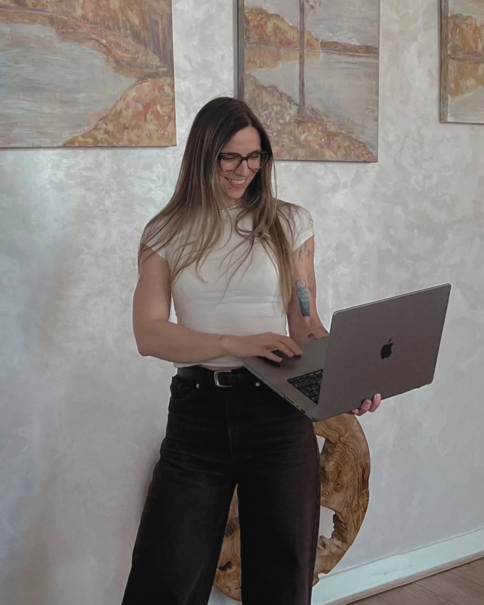 A woman with long hair, glasses, and tattoos on her arm, standing indoors, smiling while looking at a silver MacBook laptop.
