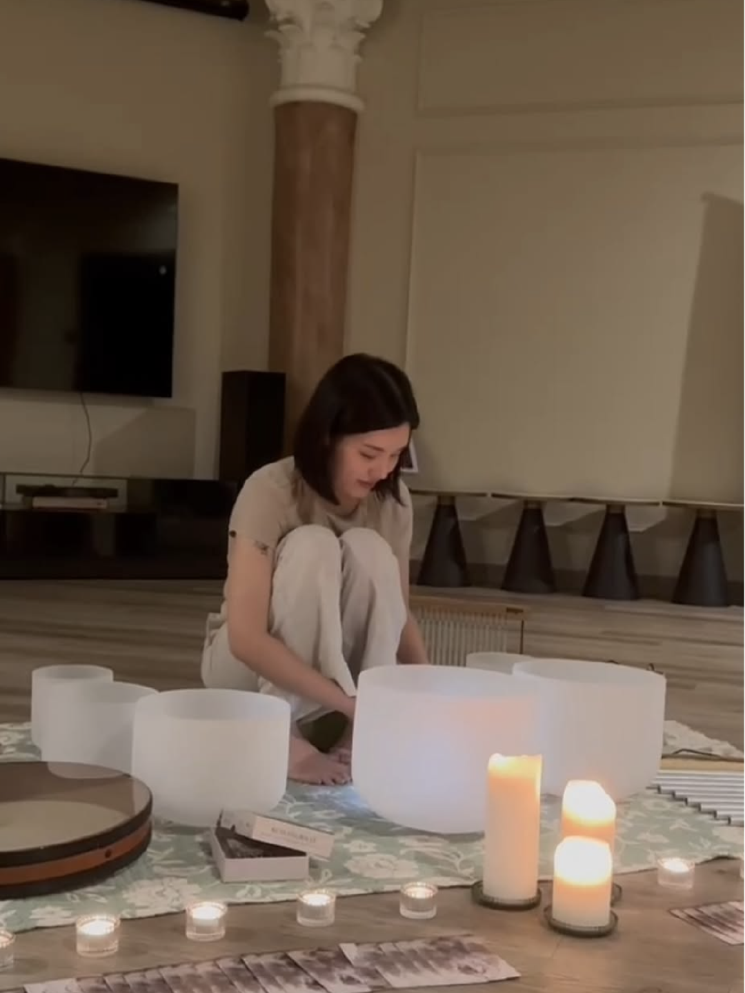 Woman sitting cross-legged on floor surrounded by candles and musical instruments, in a cozy indoor setting.
