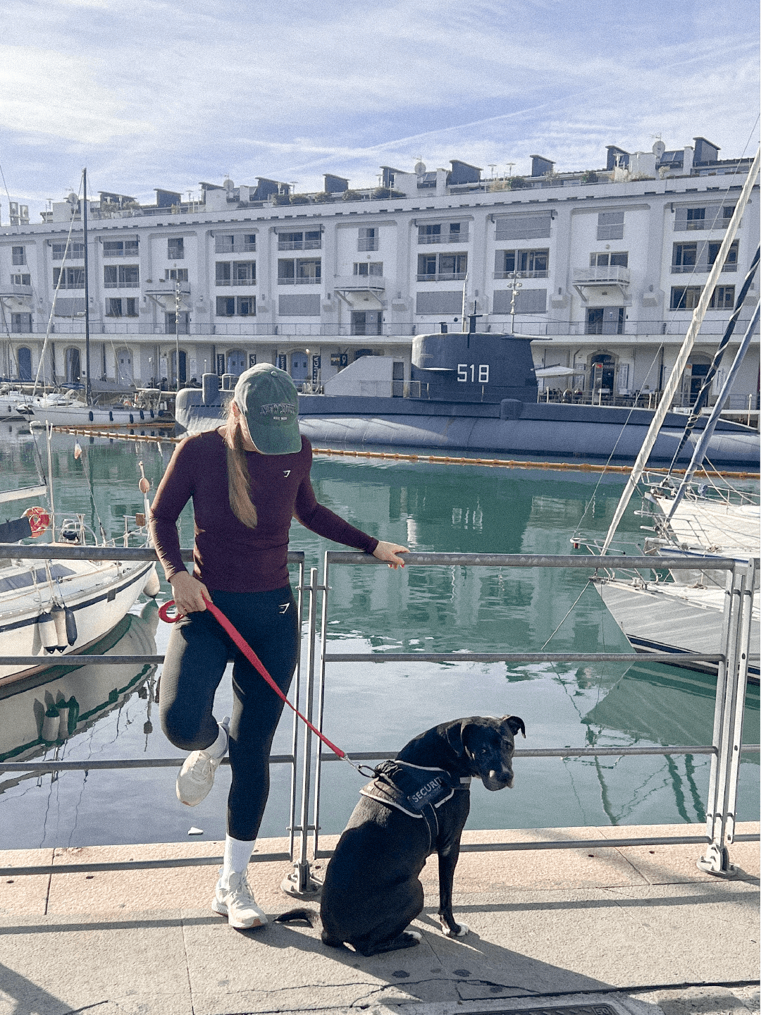 A woman in athletic clothing and a green cap holds a red leash attached to a black security dog sitting by a marina. There are boats and a large gray submarine docked in the background.