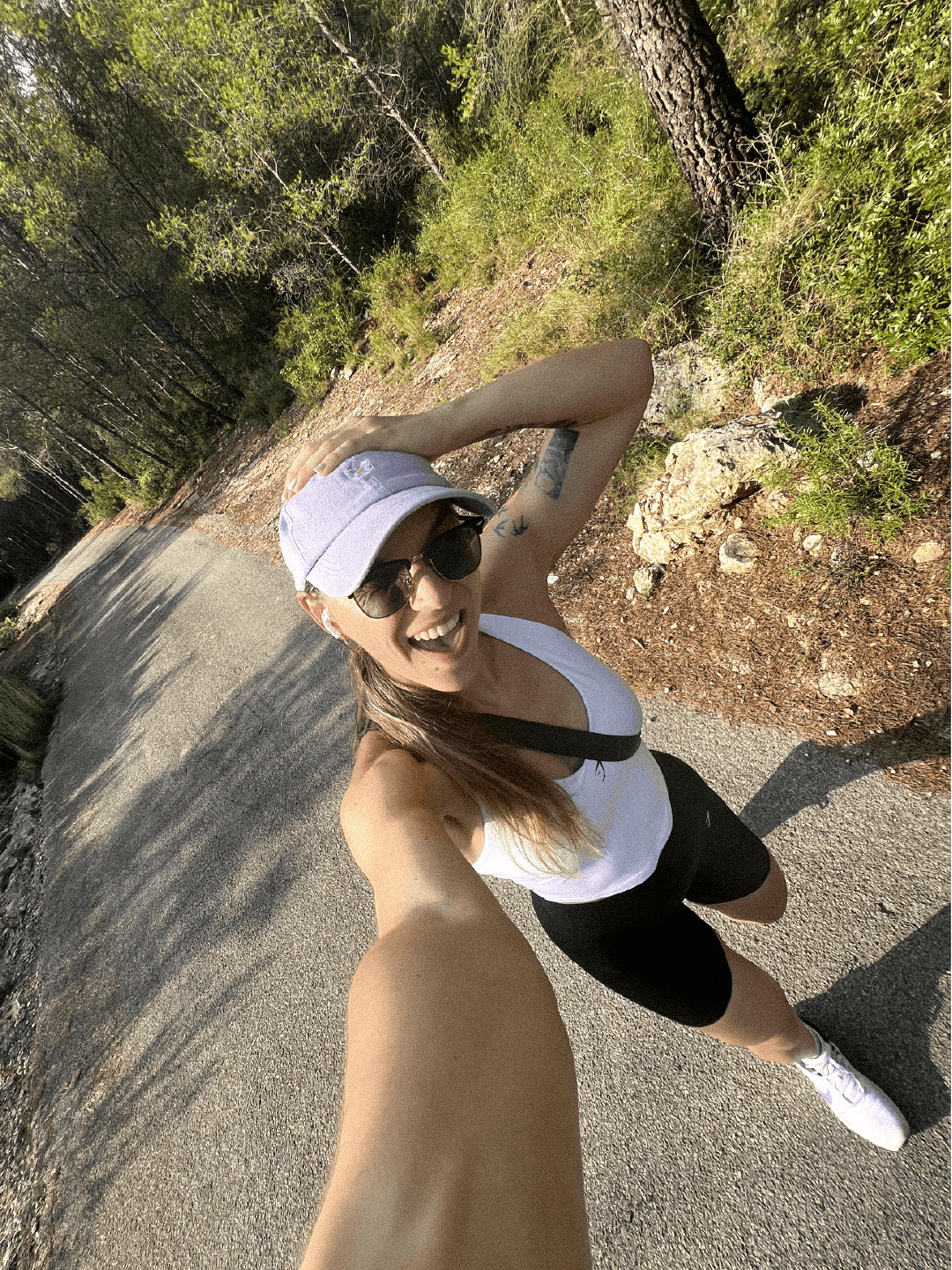 A woman taking a selfie on a trail in a forest, wearing a white tank top, black shorts, white sneakers, sunglasses, and a light purple cap, smiling cheerfully.
