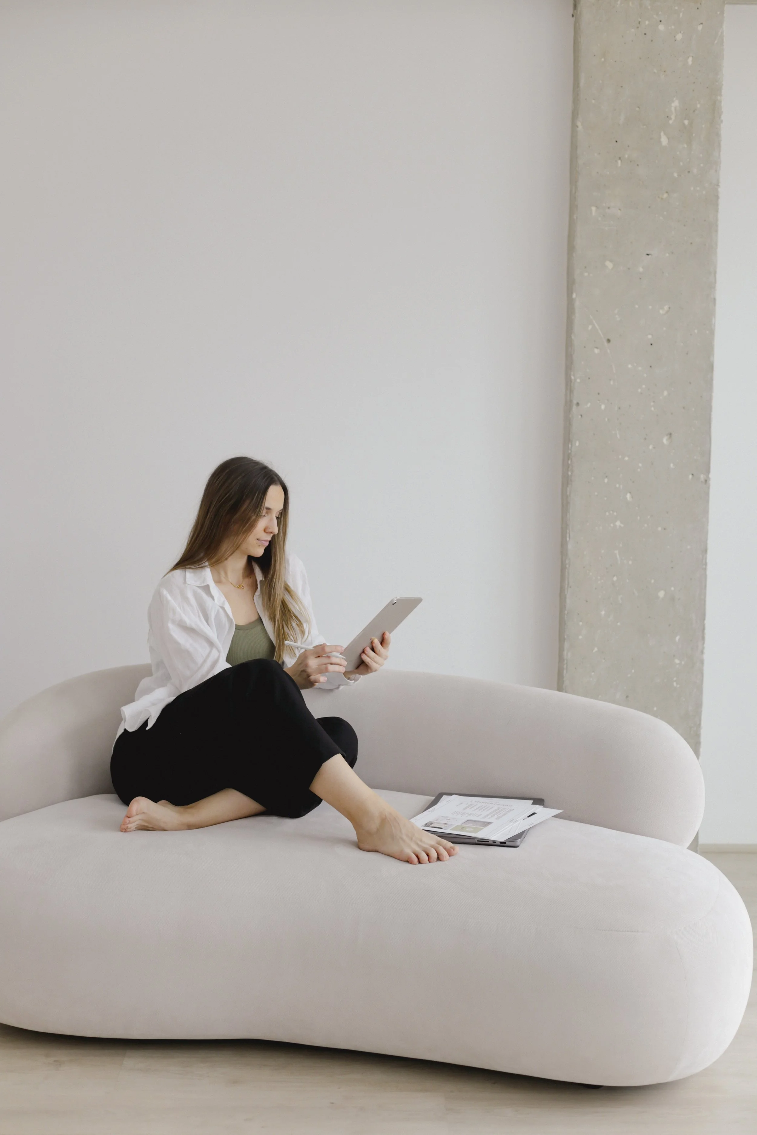 A woman sitting on a light-colored sofa, looking at a tablet in her hands, with papers on the sofa beside her in a minimalistic room.