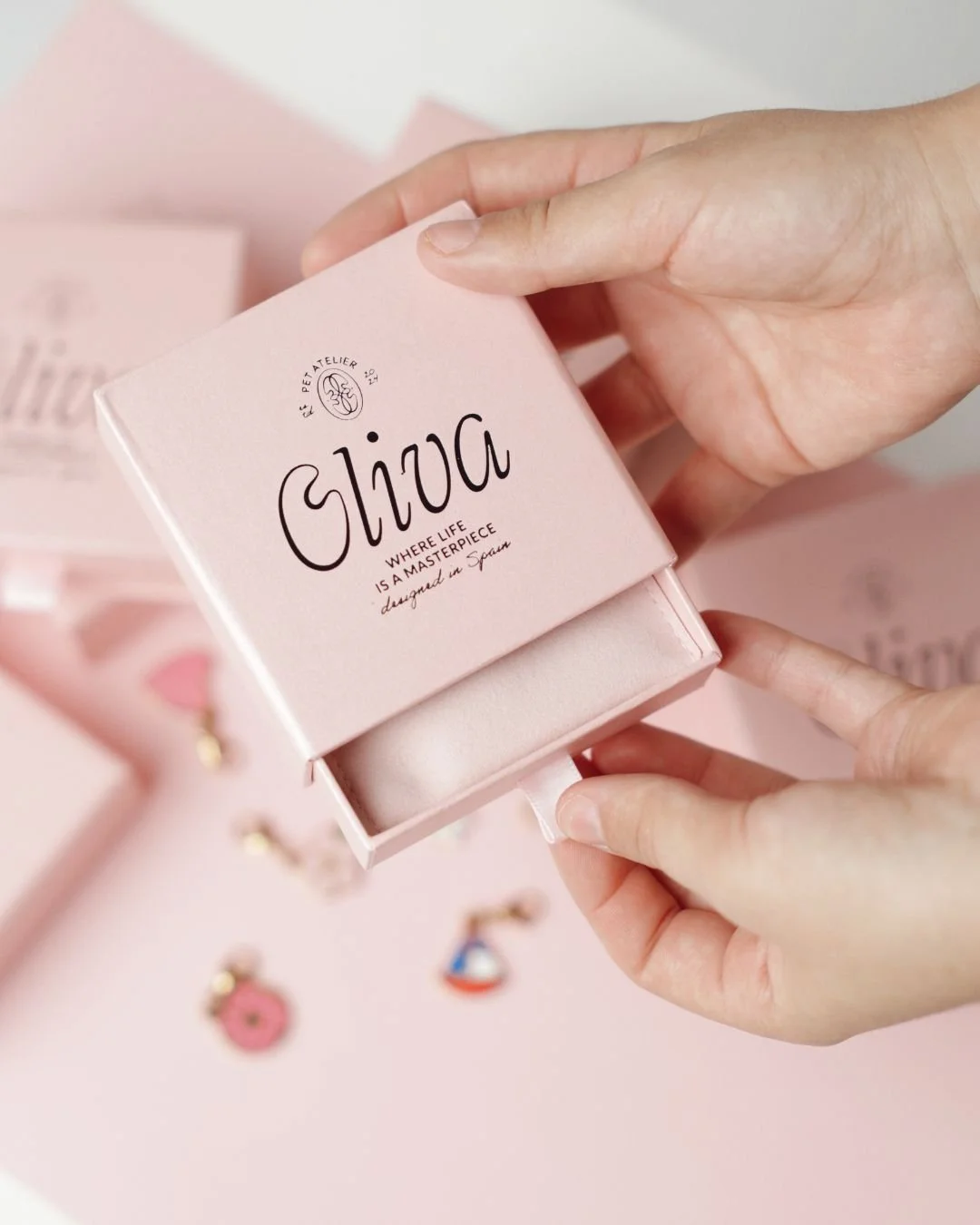 A person holding a small pink jewelry box with black text that says, "Oliiza" and "Where life is a masterpiece designed in Spain". The box is open, revealing a beige jewelry pouch inside. Several colorful earrings are blurred in the background on a p