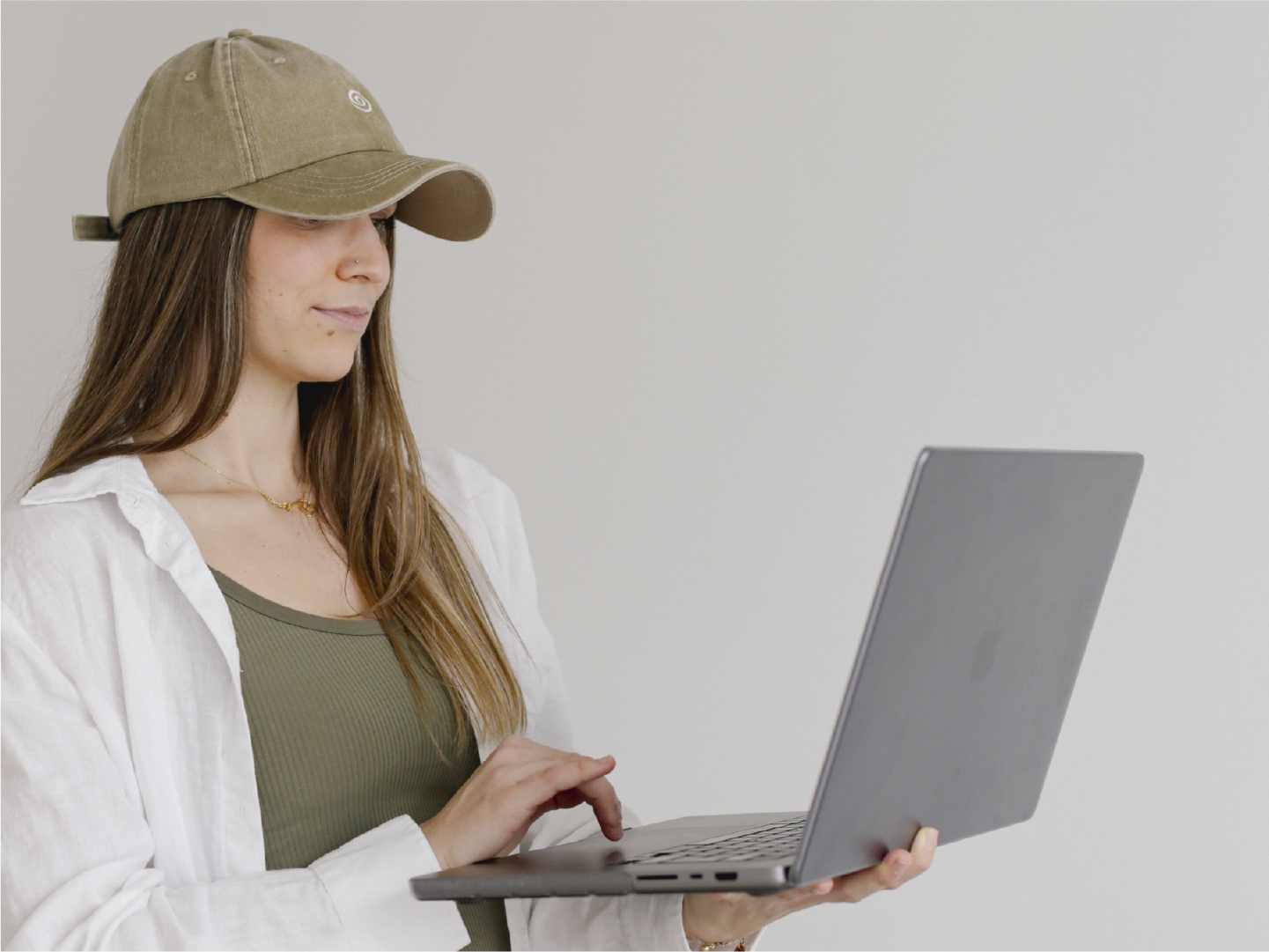 Young woman wearing a beige cap, white shirt, and green top working on a silver laptop against a plain white background.