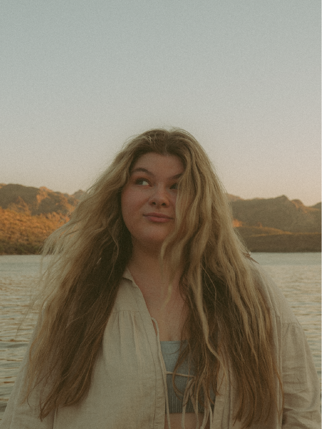 A woman with long wavy hair standing near a body of water with mountains in the background during sunset.