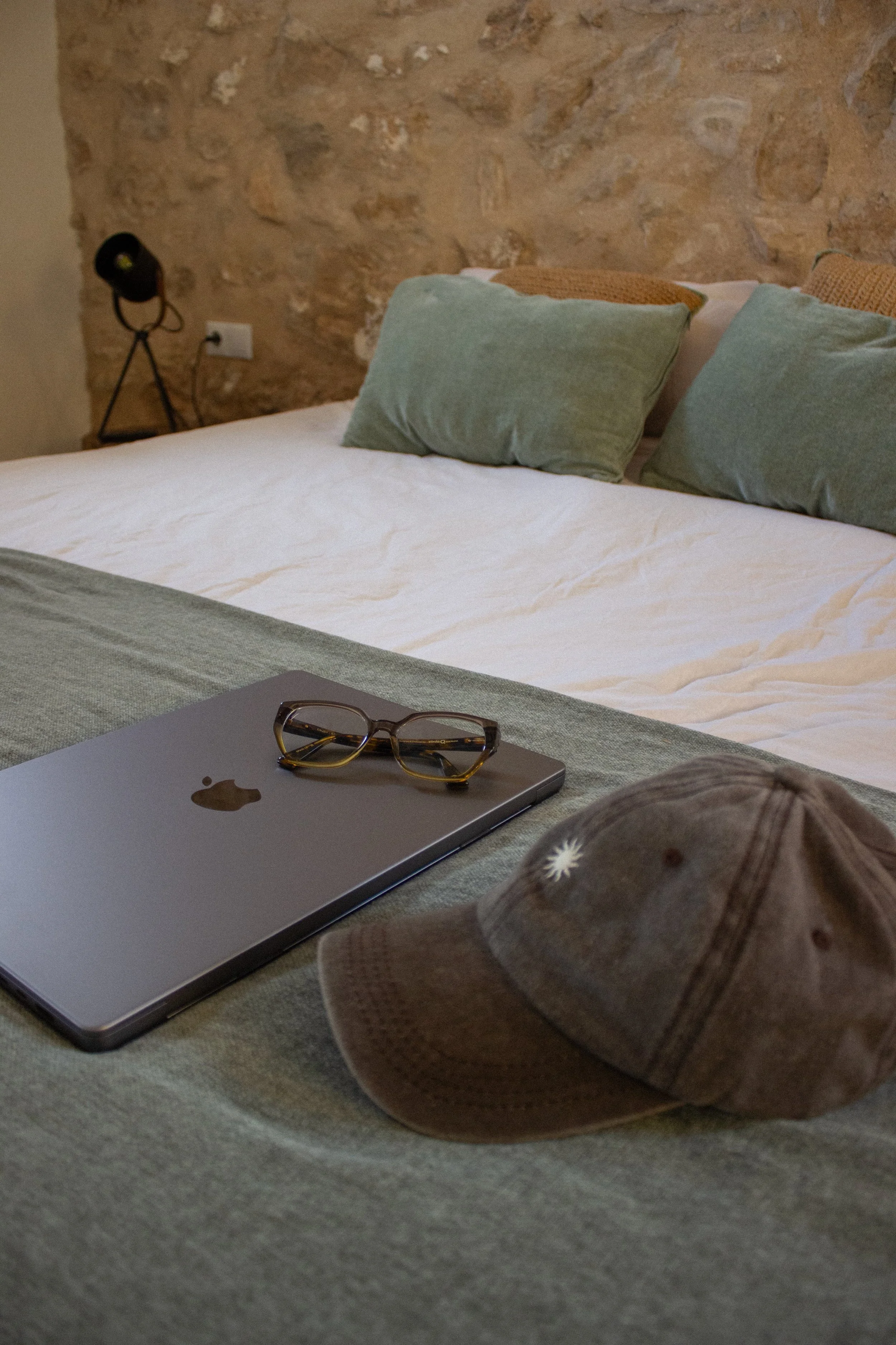 A neatly made bed with green pillows against a textured stone wall. On the bed, there is a closed silver MacBook with sunglasses on top and a brown baseball cap nearby. To the left, a modern black lamp is mounted on the wall, plugged into an electrical outlet.