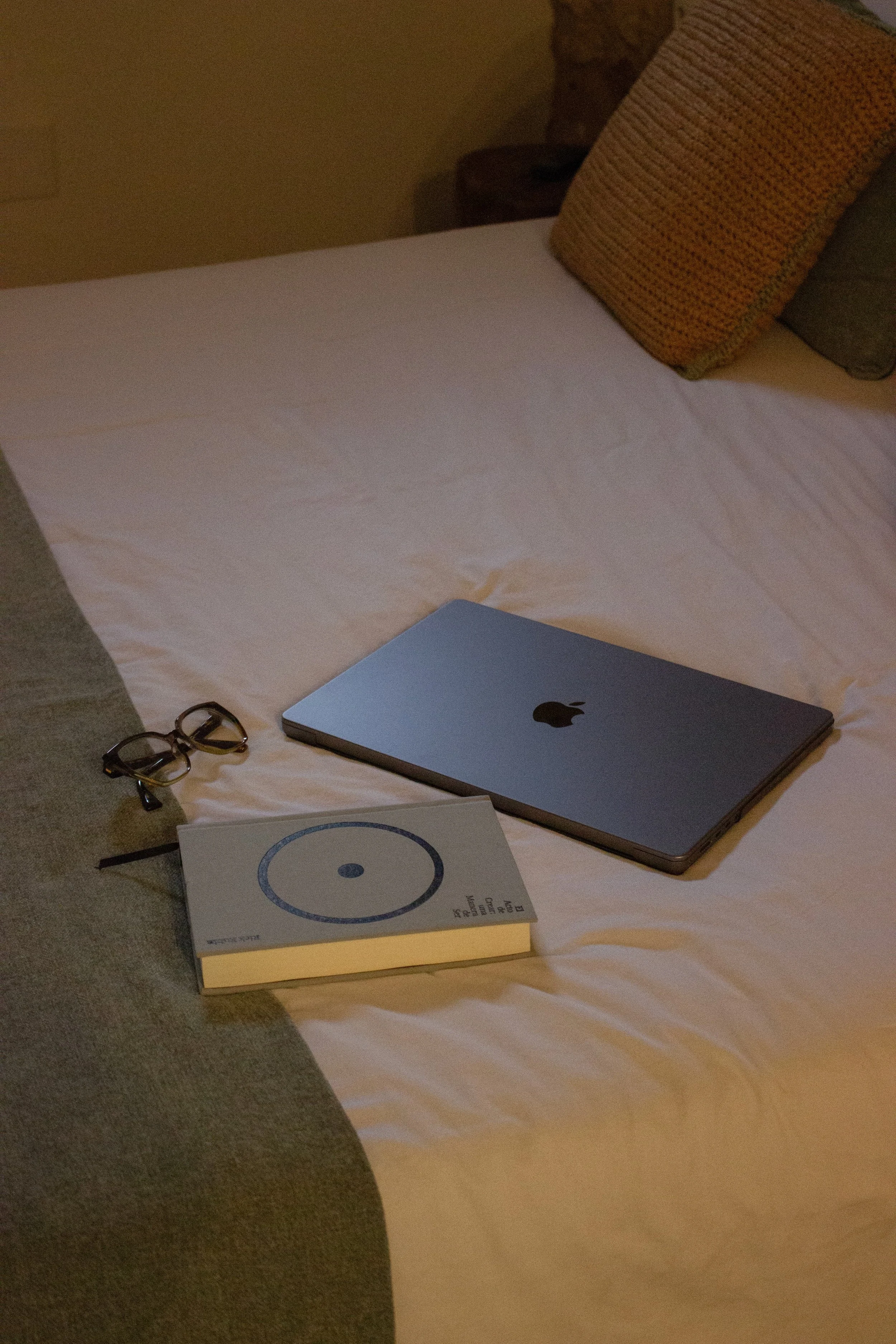 A bedroom with a bed covered in white sheets, a closed silver MacBook, a pair of glasses, and a book on the cover placed on the bed. There are pillows in beige and gray colors, a nightstand, and a wall in the background.