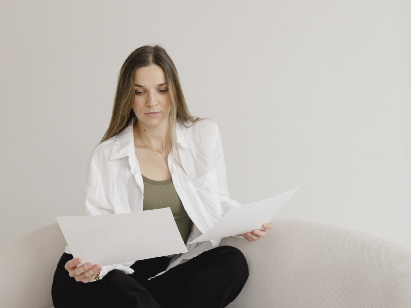 A woman with long light brown hair sits on a light-colored sofa, examining two sheets of white paper, wearing a white shirt over a green top and black pants.