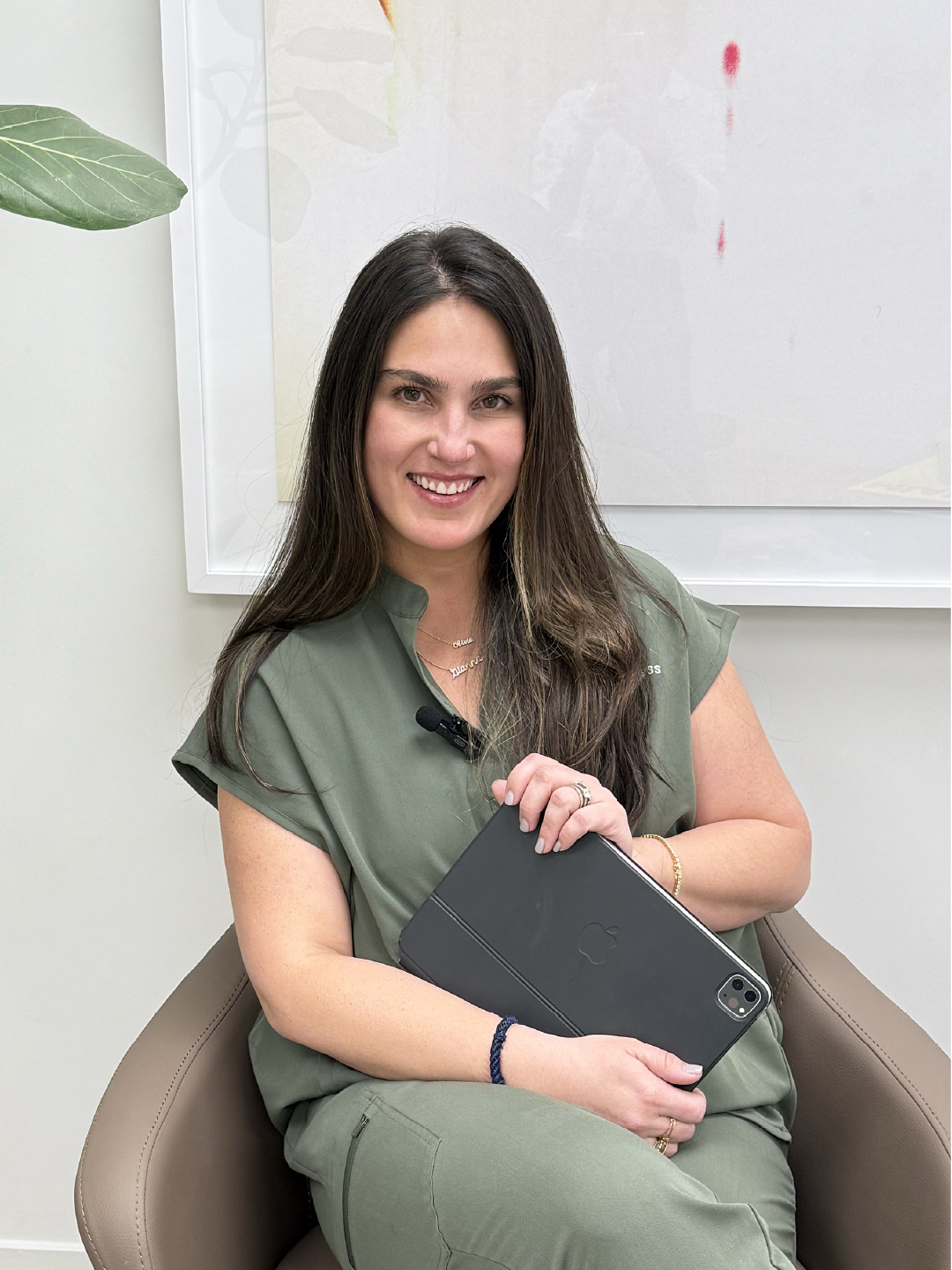 A woman with long brown hair, smiling, sitting in a beige chair with a black Apple iPad in her hands, in an office setting with a whiteboard behind her and a green plant nearby.