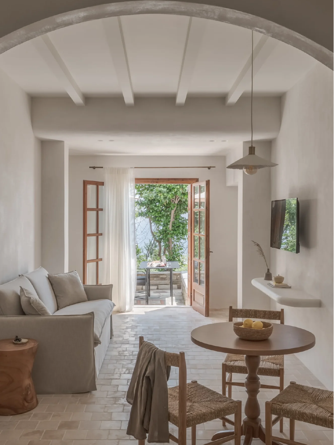 Living room with white walls, a beige sofa, small wooden side table, round wooden dining table with three chairs, a wall-mounted TV, and open double doors leading to a balcony with greenery.