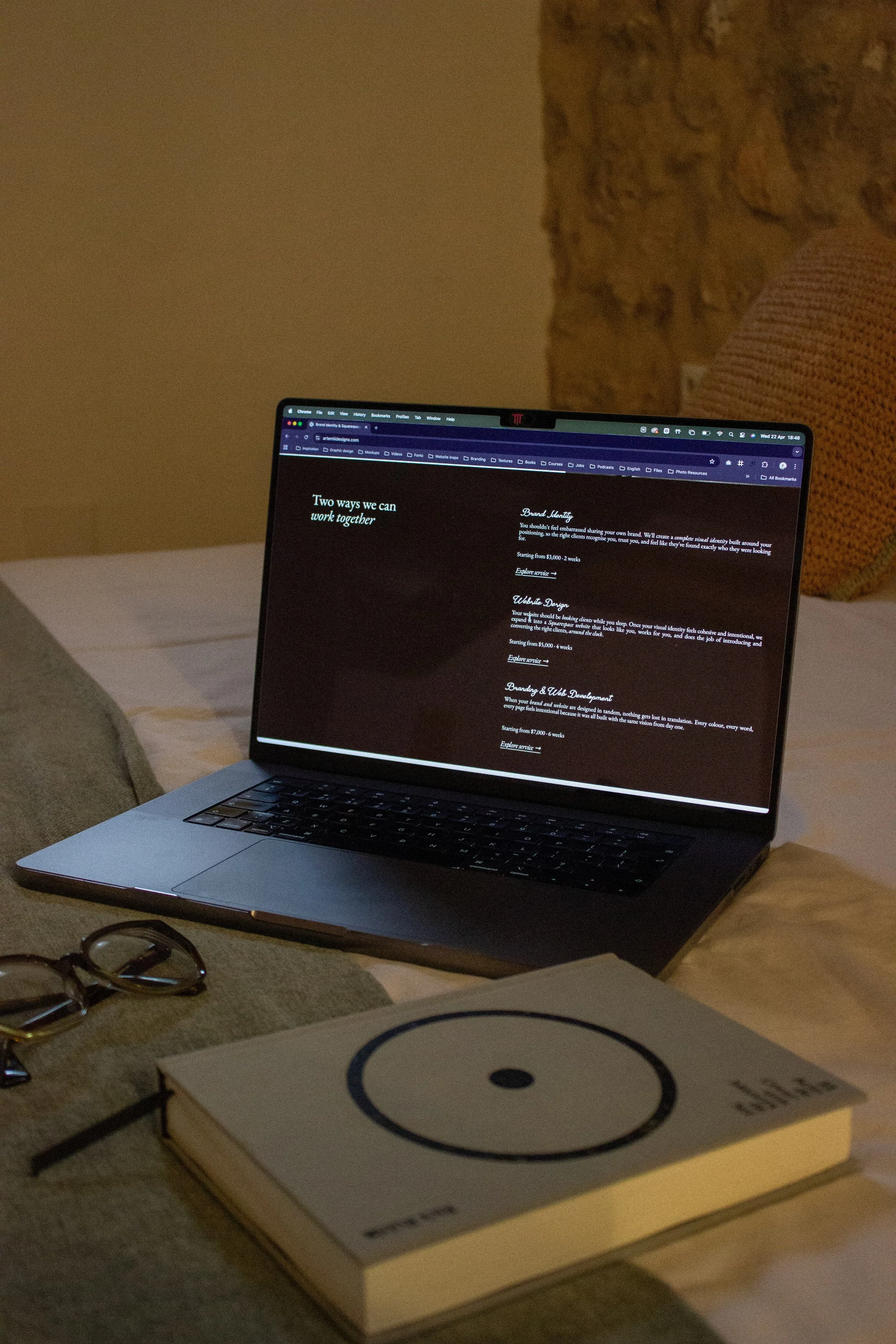 A laptop displaying a Artentii Designs website services, with a book, a pair of glasses, and a notebook nearby. On the screen you can see her website and branding services she offers for women-led brands.