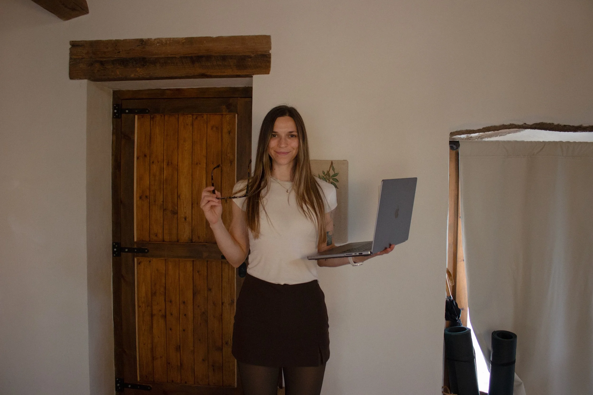 A brand identity and Squarespace designer founder of Artentii Designs. She has long brown hair, dressed in a white top and black skirt, standing in a room holding a laptop in one hand and glasses in the other.