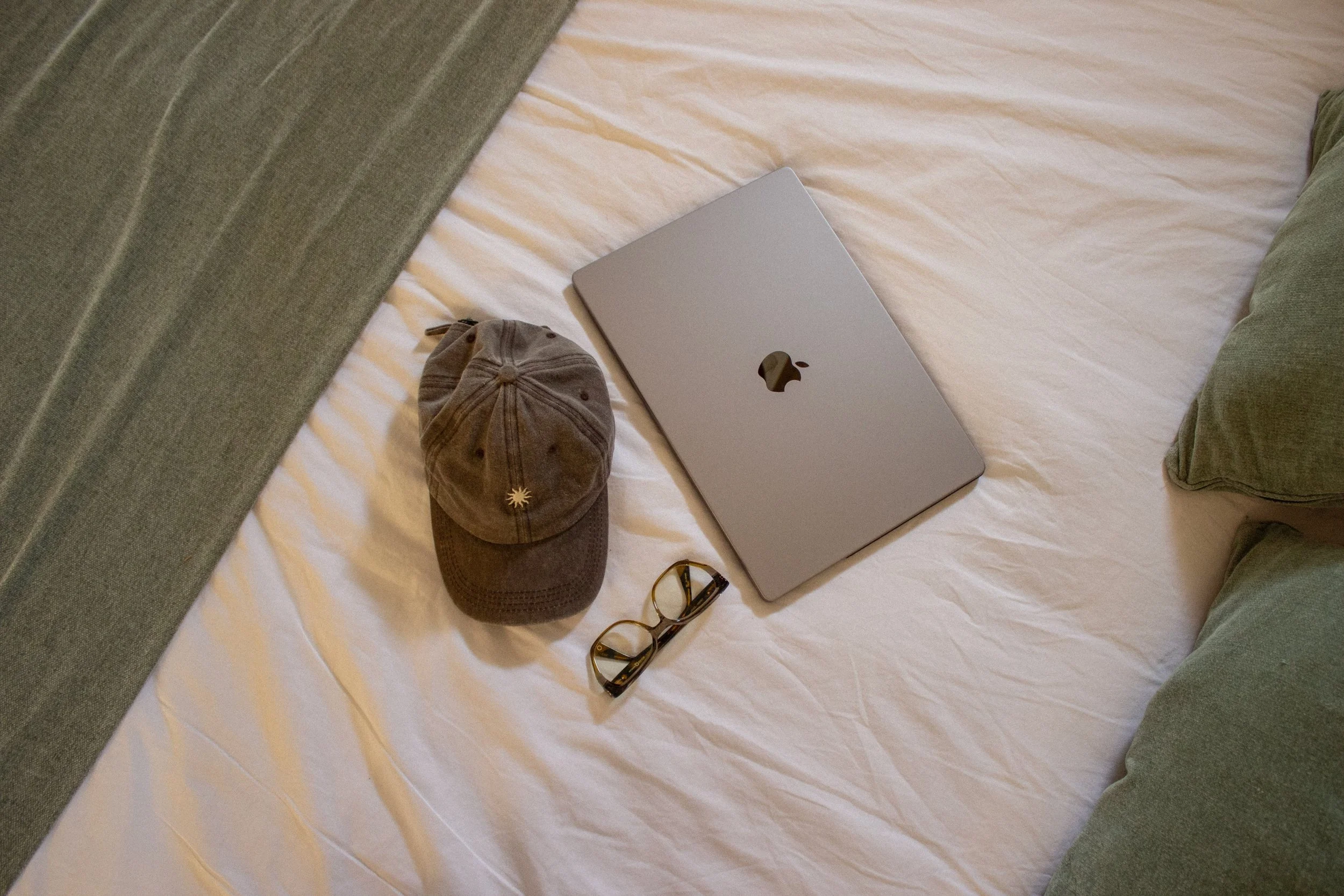 A neatly made bed with a closed silver MacBook laptop, a brown baseball cap, a pair of glasses, and a black and yellow wristwatch on it. The bed has cream-colored sheets, and there are green pillows and a gray headboard visible.