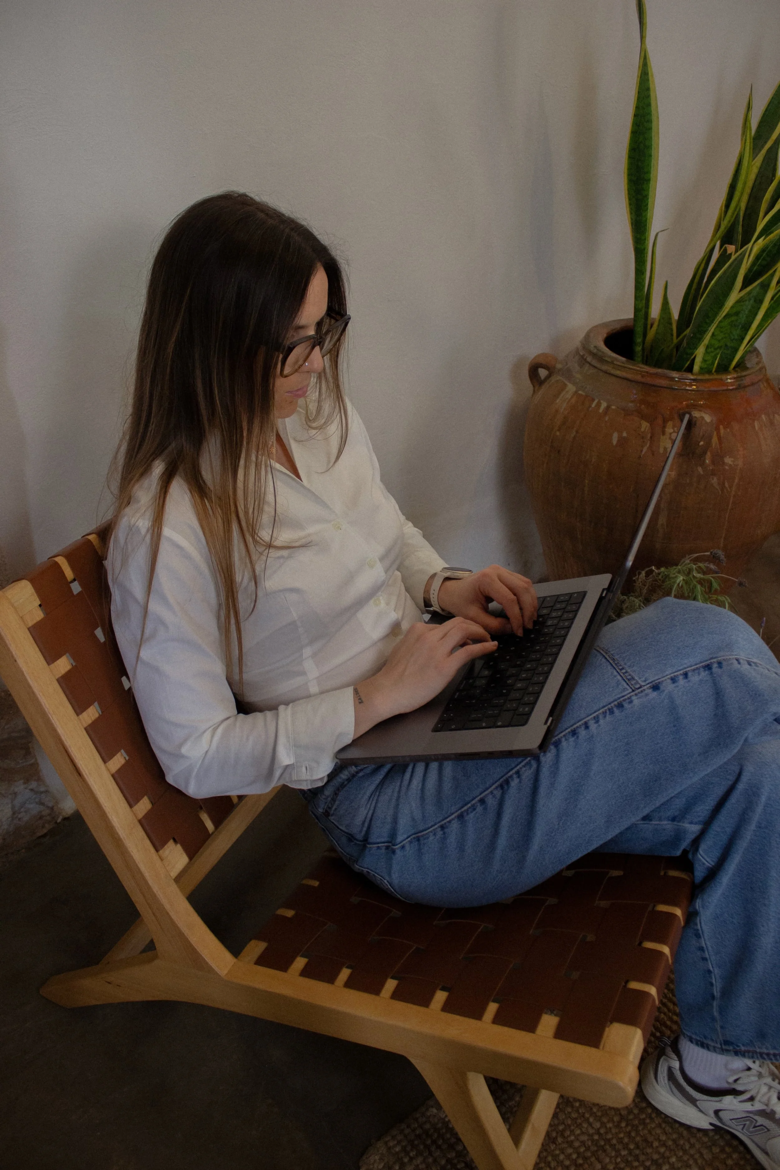 Brand Identity and Squarespace designer Marina Terentii, founder of Artentii Designs. Marina wears glasses and is sitting on a wooden chair, using a laptop in a room with a large ceramic pot with green plants nearby.