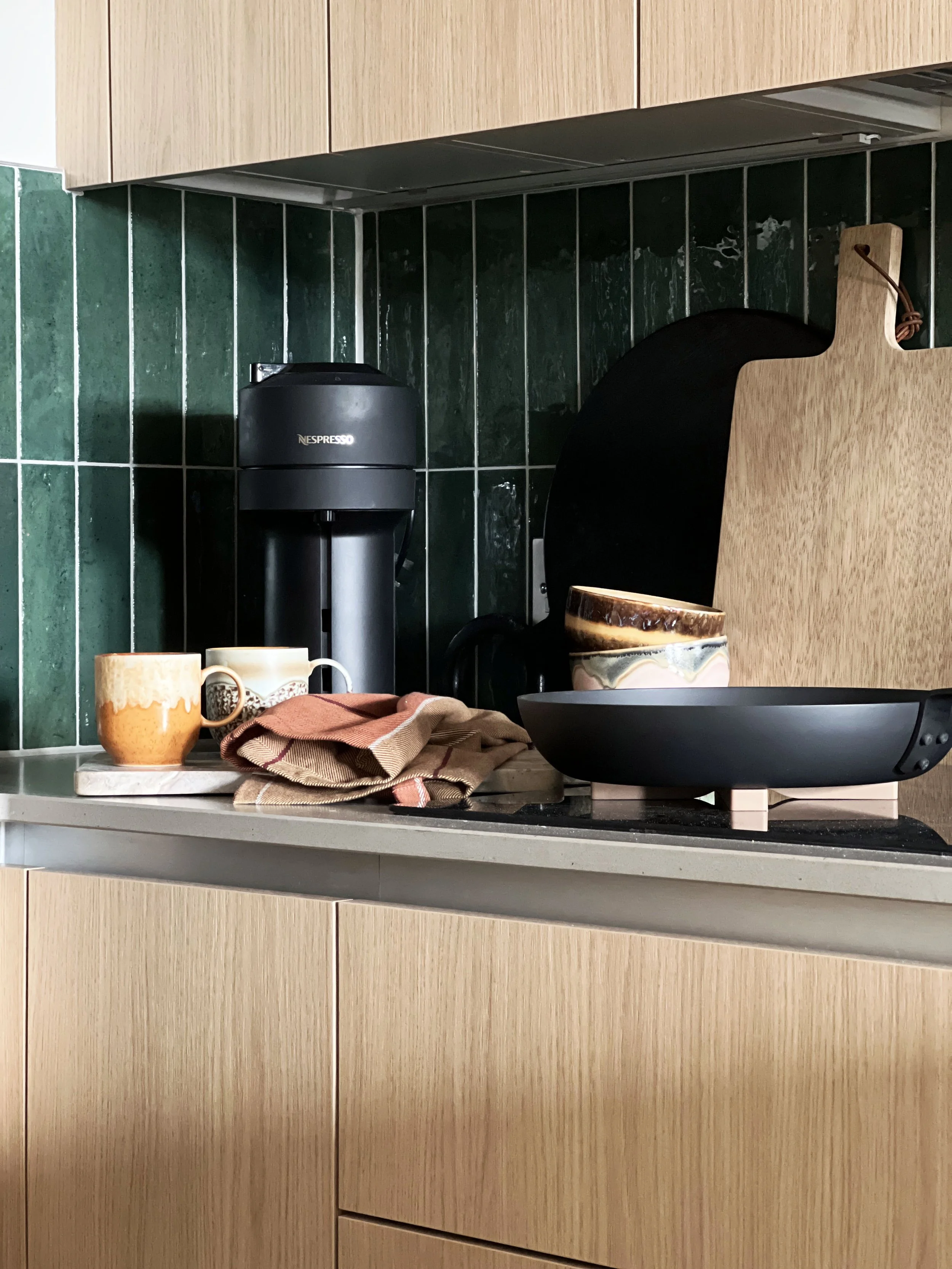 An oak kitchen with dark green tiles.  A black frying pan on the hob and glazed ceramic mugs