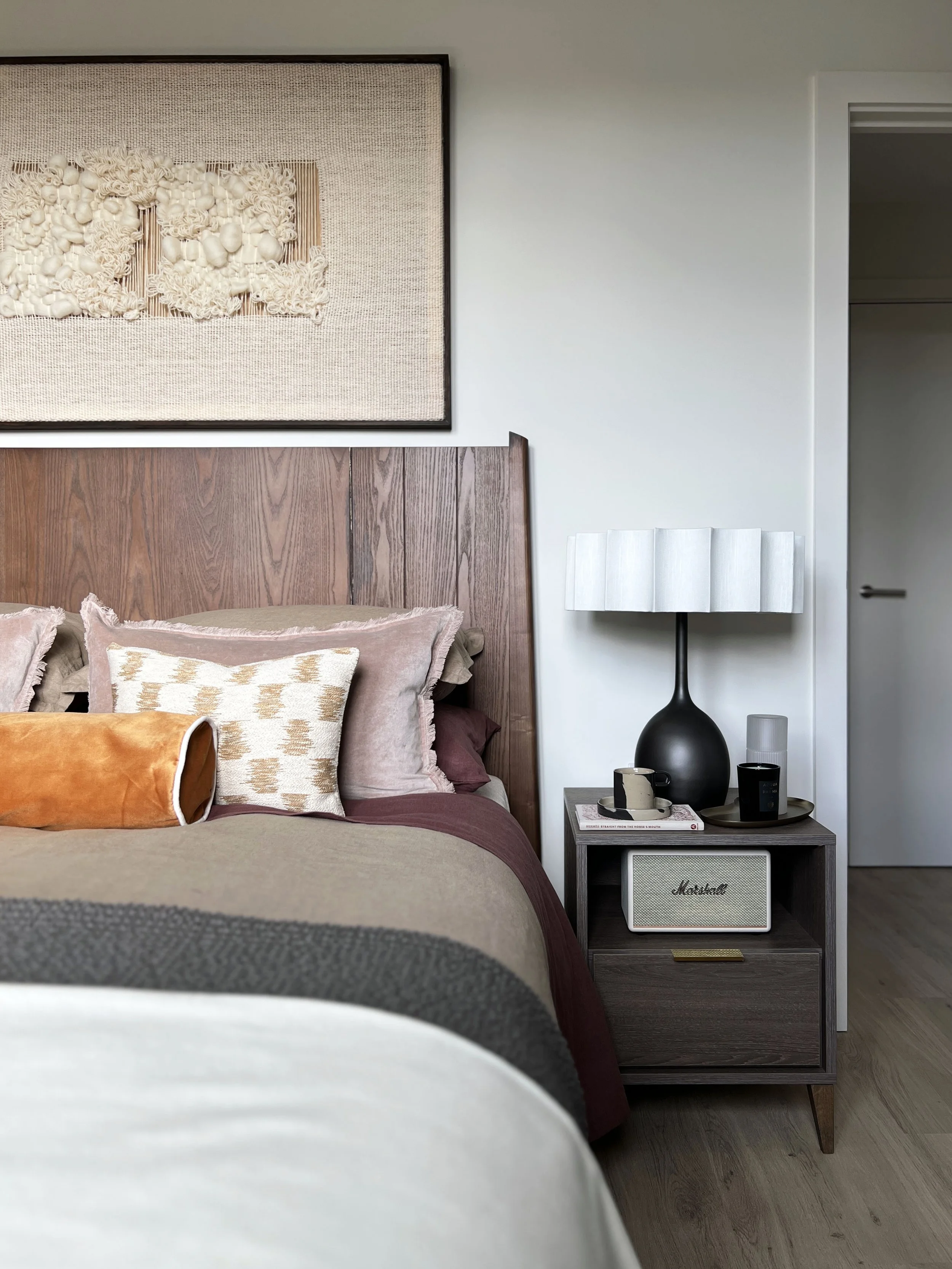 Close up of a wooden bed frame and bedside table.  A fluted lamp is on the bedside table and some scatter cushions in orange and pink are on the bed