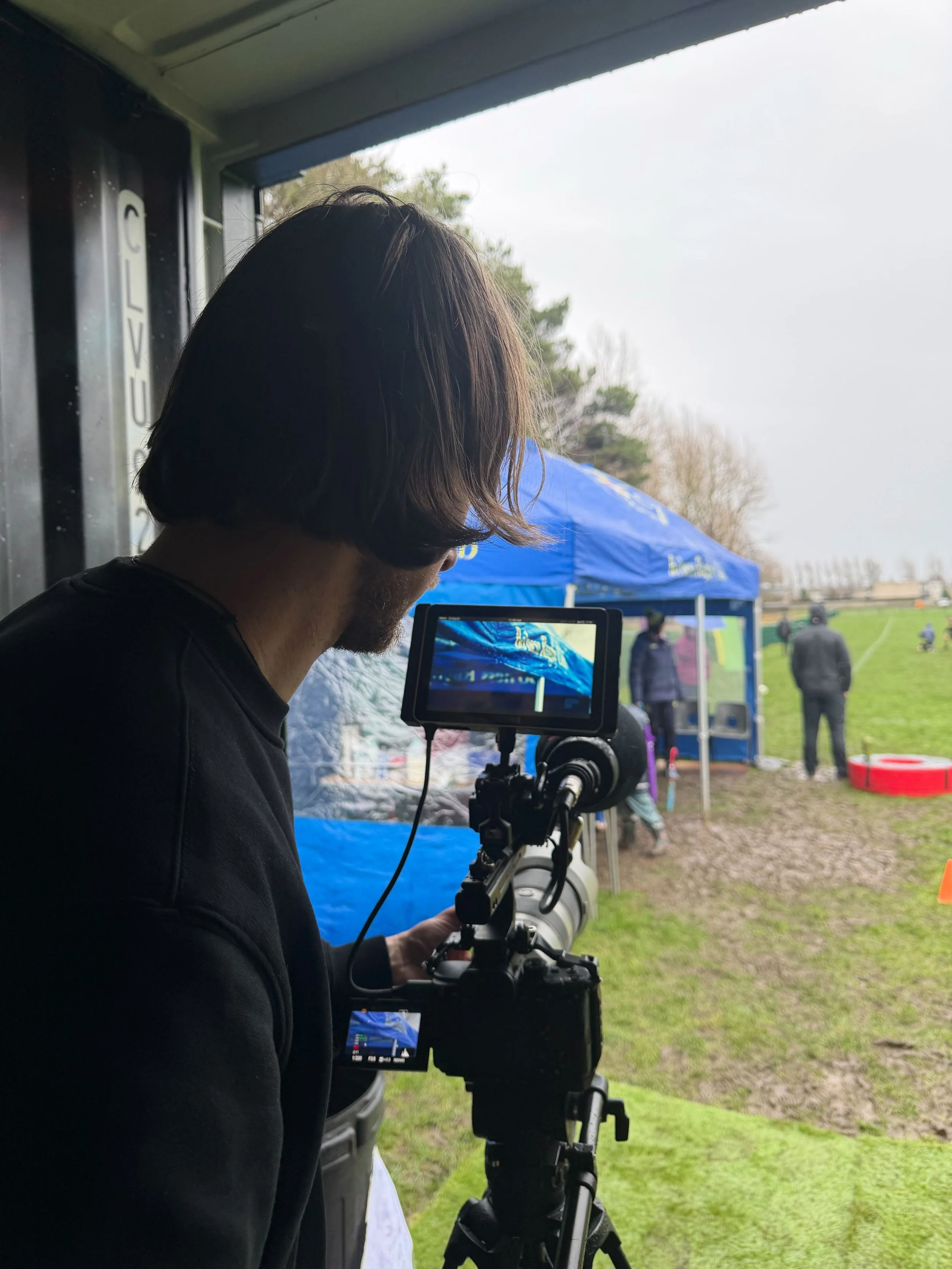 A man with shoulder-length hair and a beard operating a professional camera on a tripod, filming an outdoor event under a blue canopy tent, with people and trees in the background.