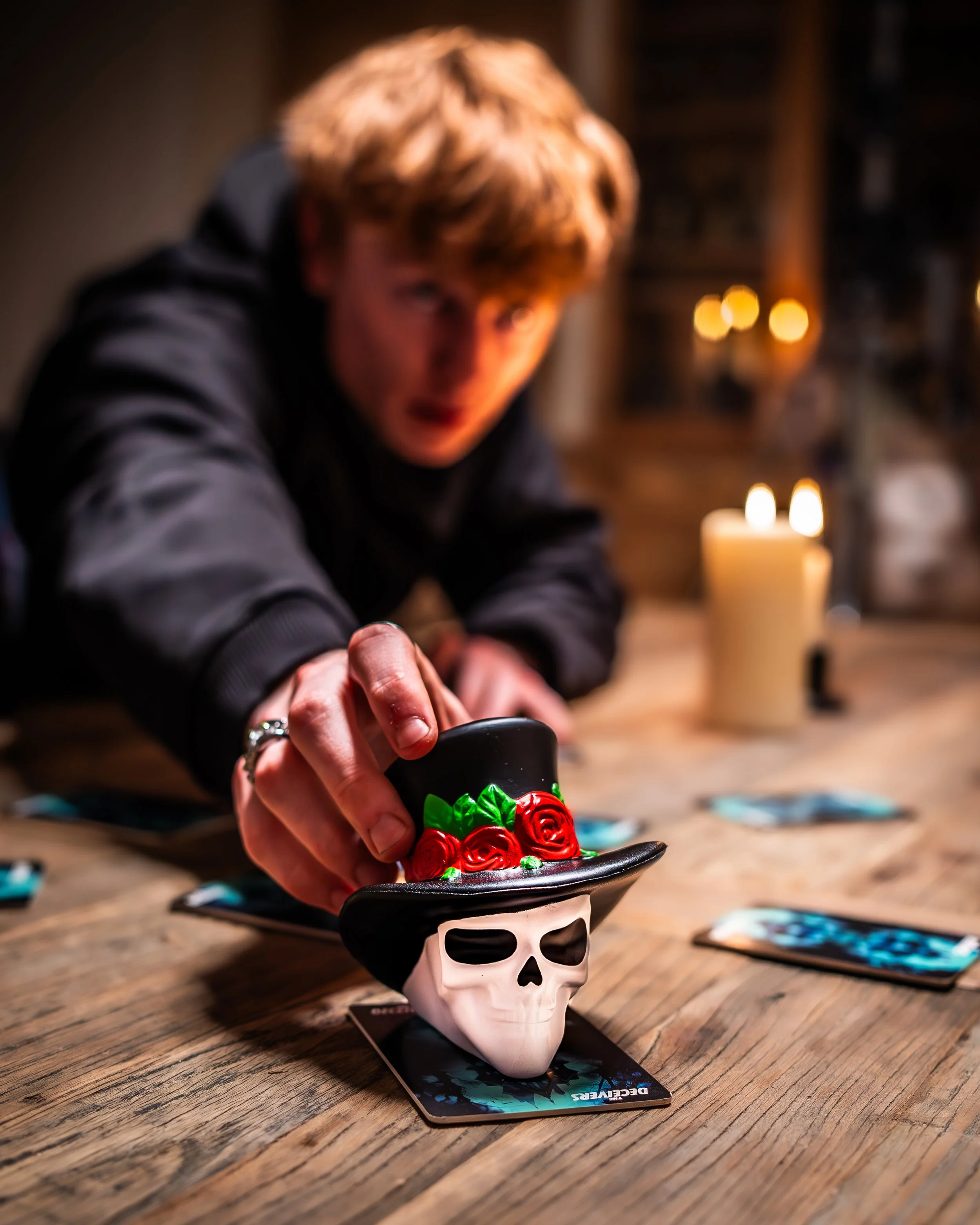 A person with reddish hair reaching out over a table decorated with video game cards and a skull-shaped candle holder with a black top hat decorated with red roses and green leaves. A flickering candle is visible in the background.