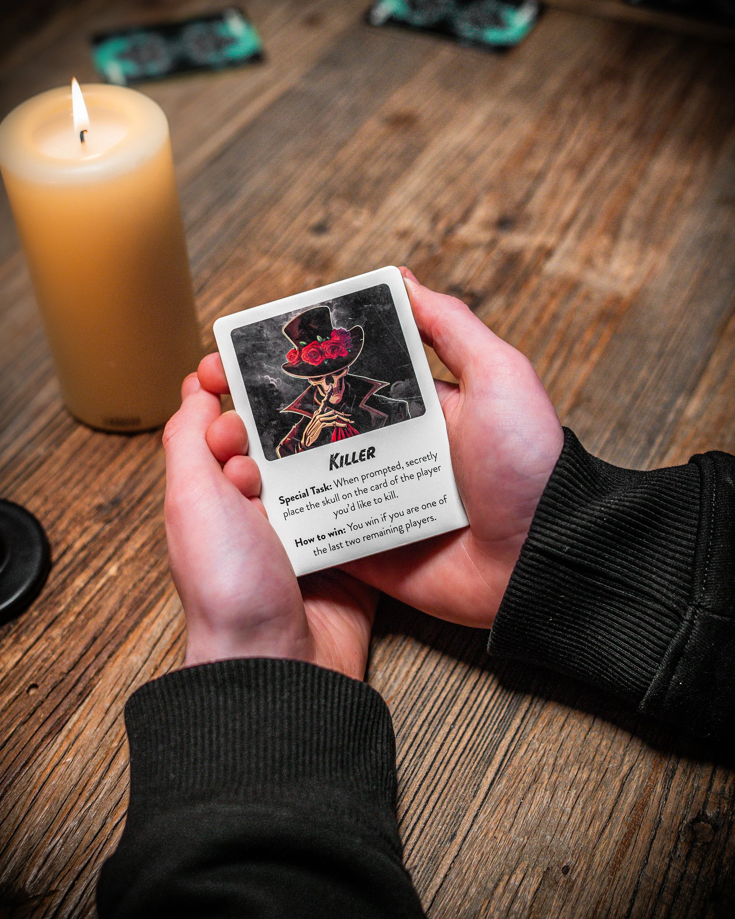 Person holding a playing card titled 'Killer' with an illustration of a skeleton wearing a top hat decorated with red roses, sitting at a wooden table near a lit beige candle.