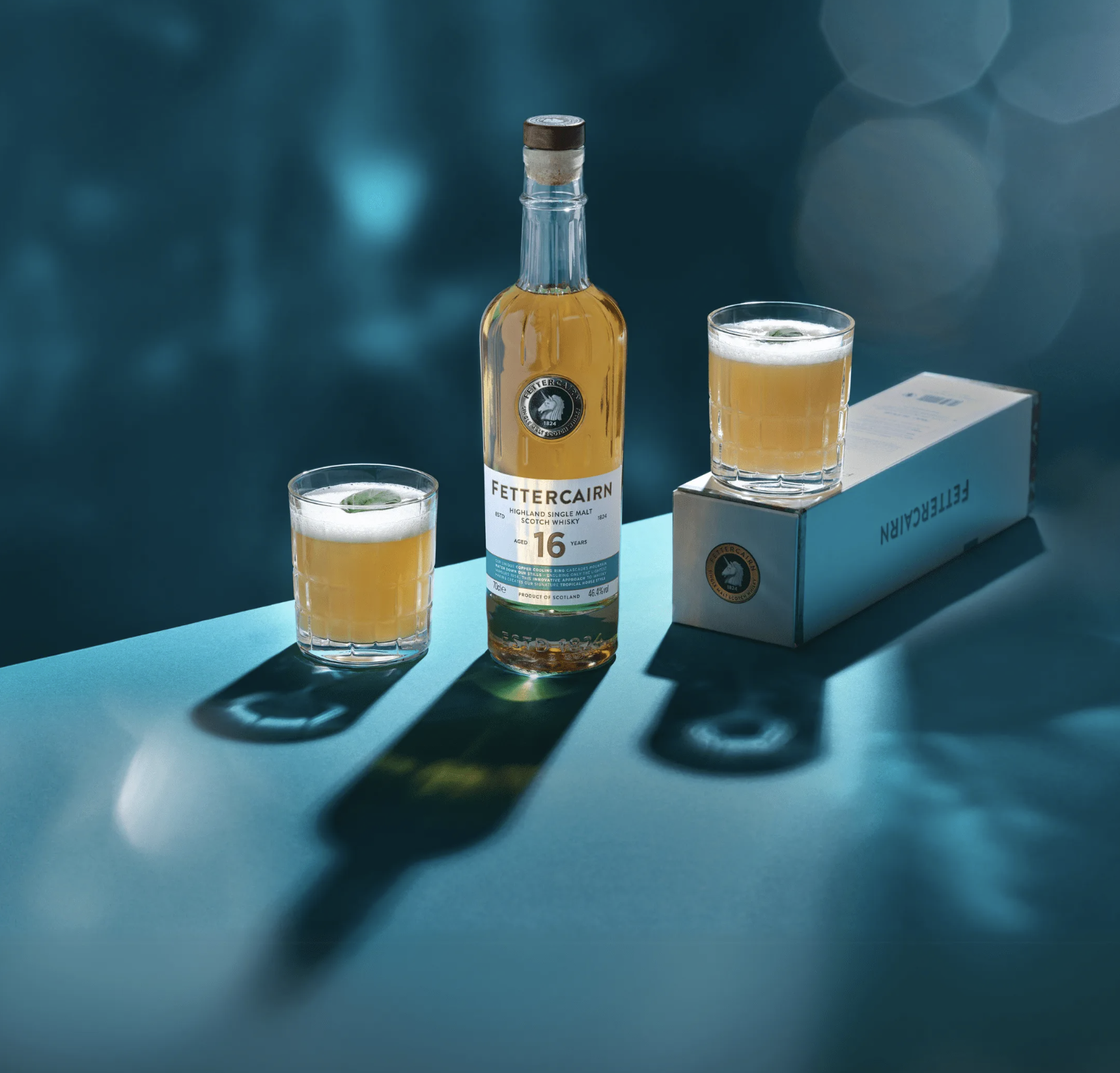 Bottle of Fettercairn Highland Single Malt Scotch Whisky with two glasses filled with whisky on a teal surface, one glass on a box labeled Fettercairn.
