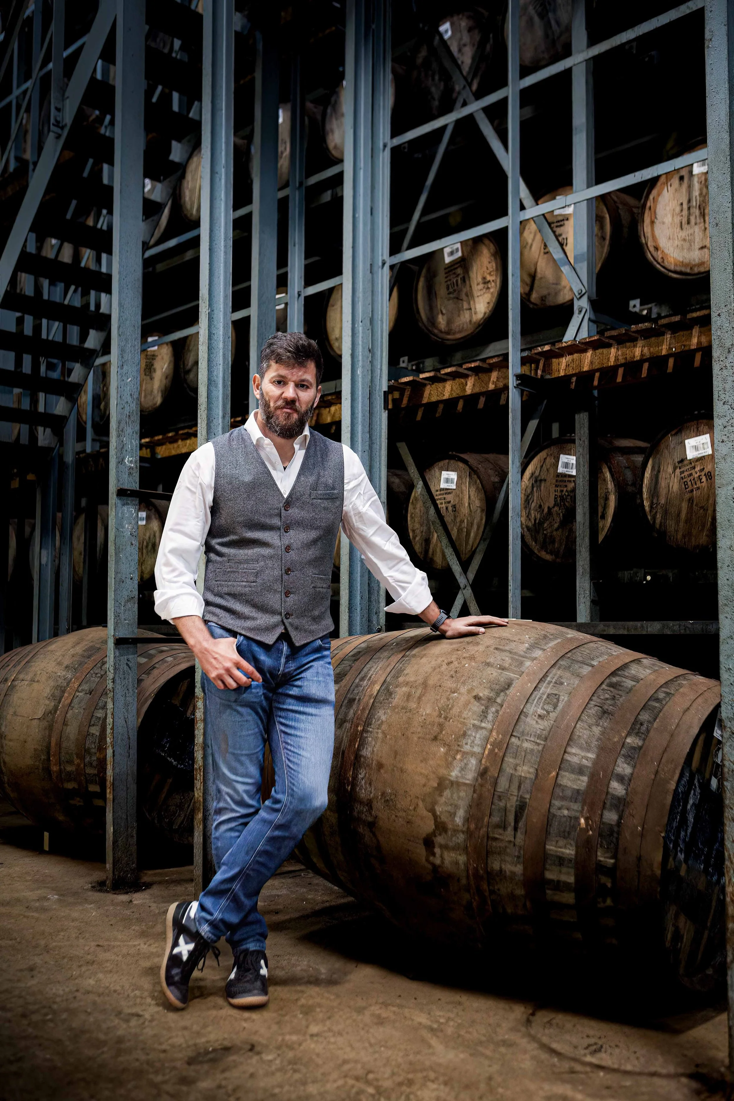 A man leaning against a large wooden whisky barrel in a warehouse with shelves of similar whisky barrels in the background.