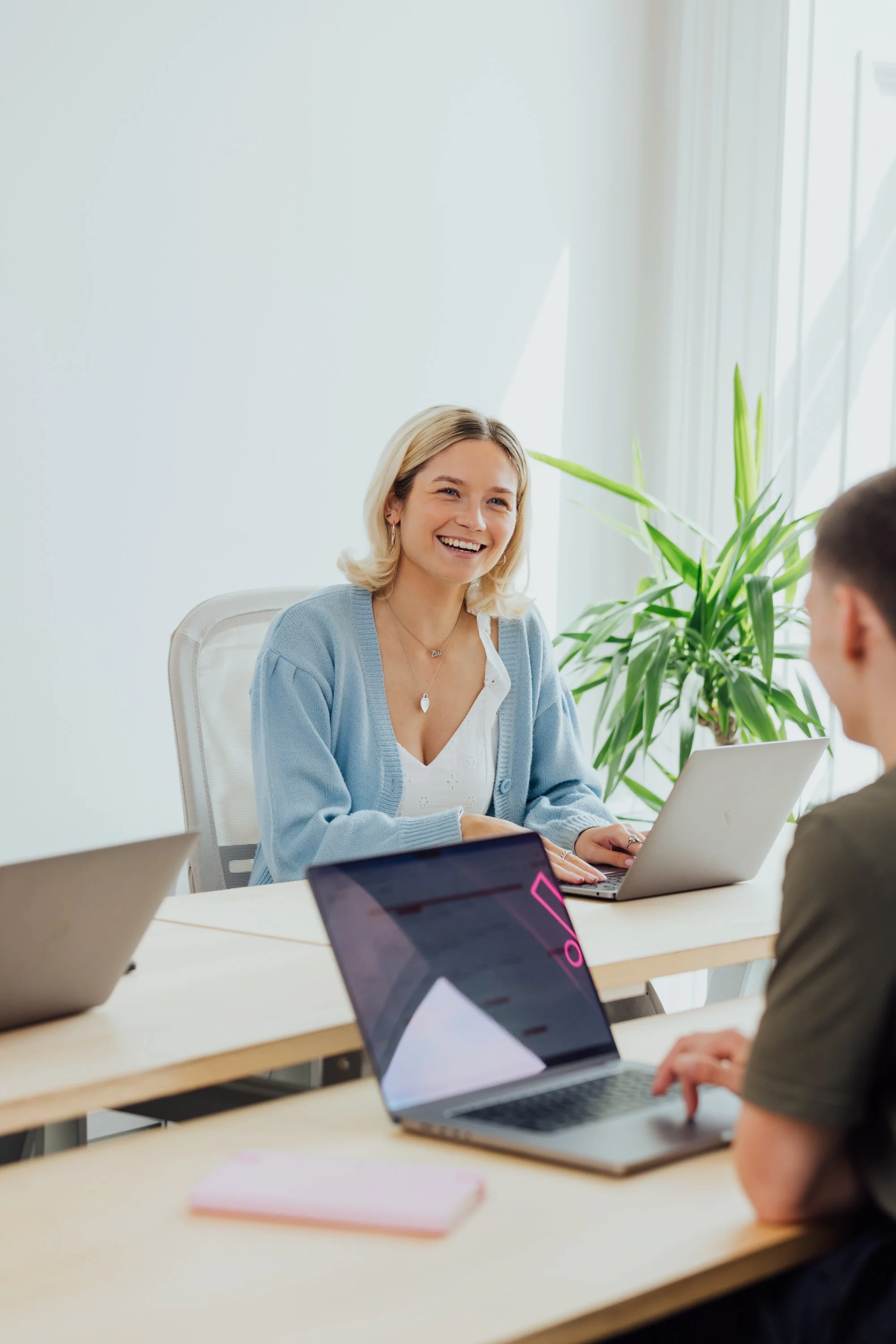 A woman smiling during a meeting with two colleagues in a bright office, using a laptop.